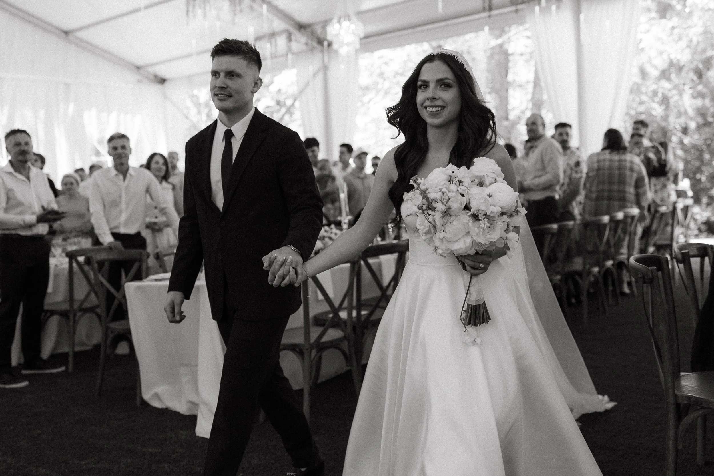 Bride and groom walk hand in hand into the reception tent as guests cheer