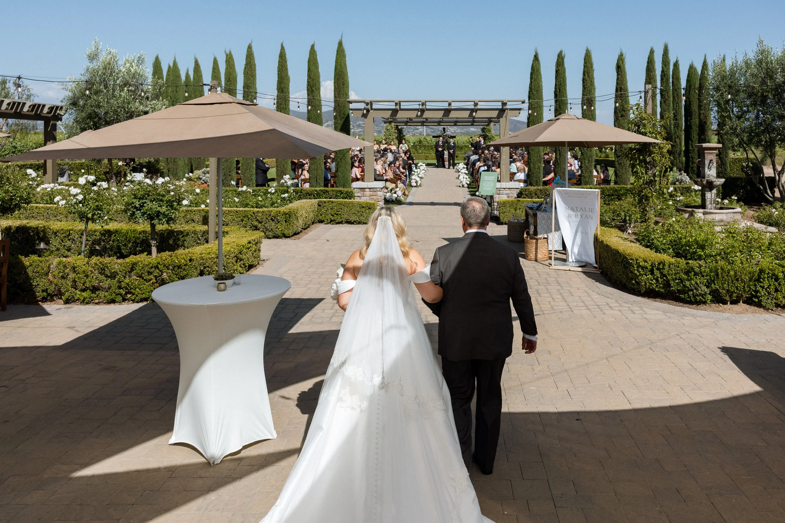 Bride and her father walk toward the ceremony aisle past a welcome sign