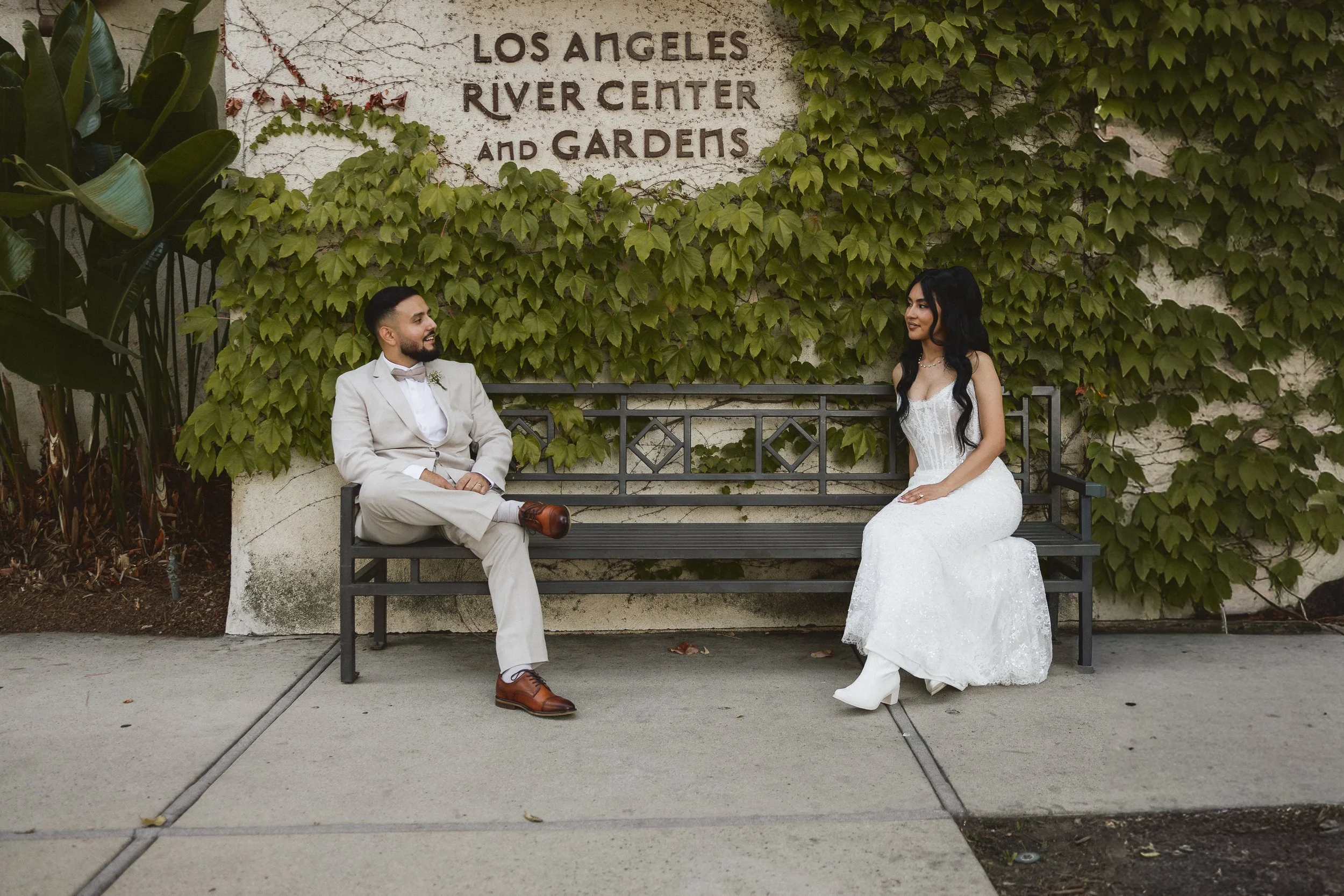 Bride and groom seated on bench with ivy wall backdrop at Los Angeles River Center and Gardens.