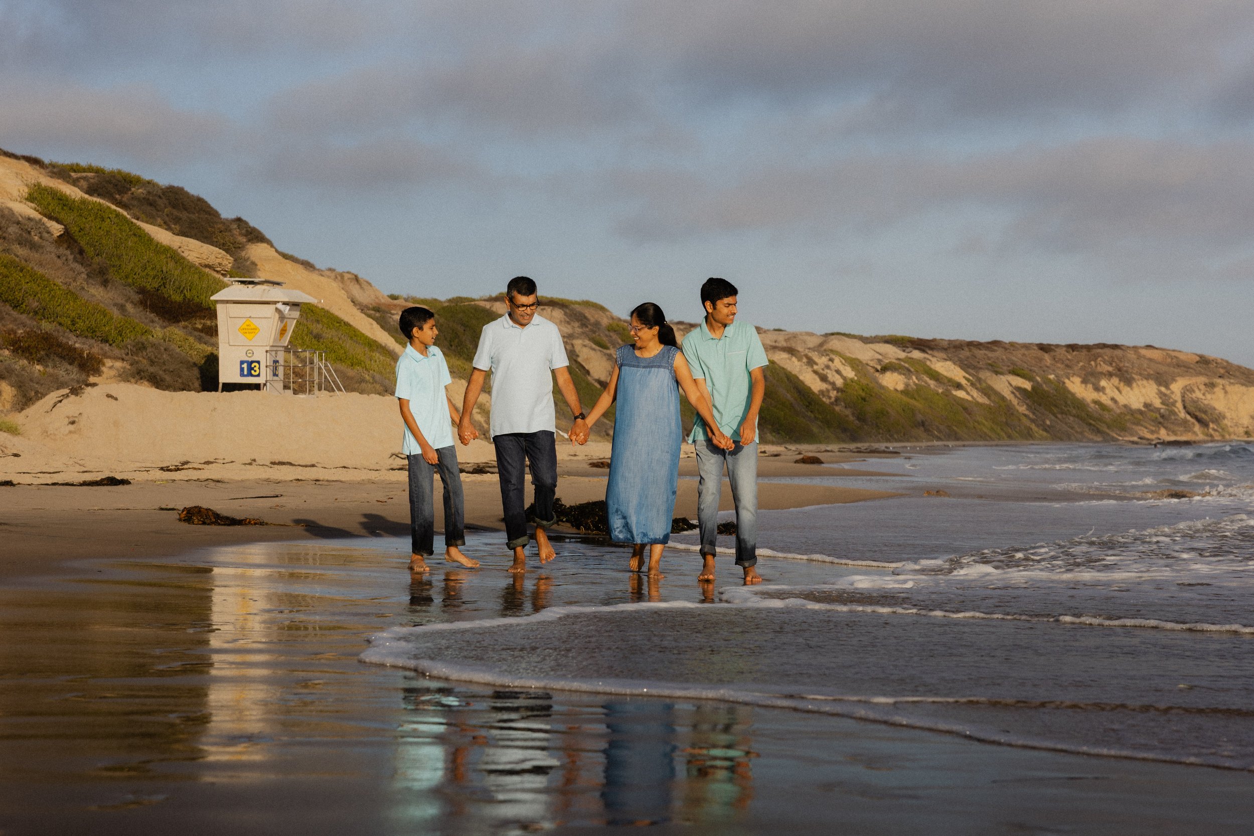Family of four holding hands and walking along the shoreline during their Crystal Cove State Park photo session.