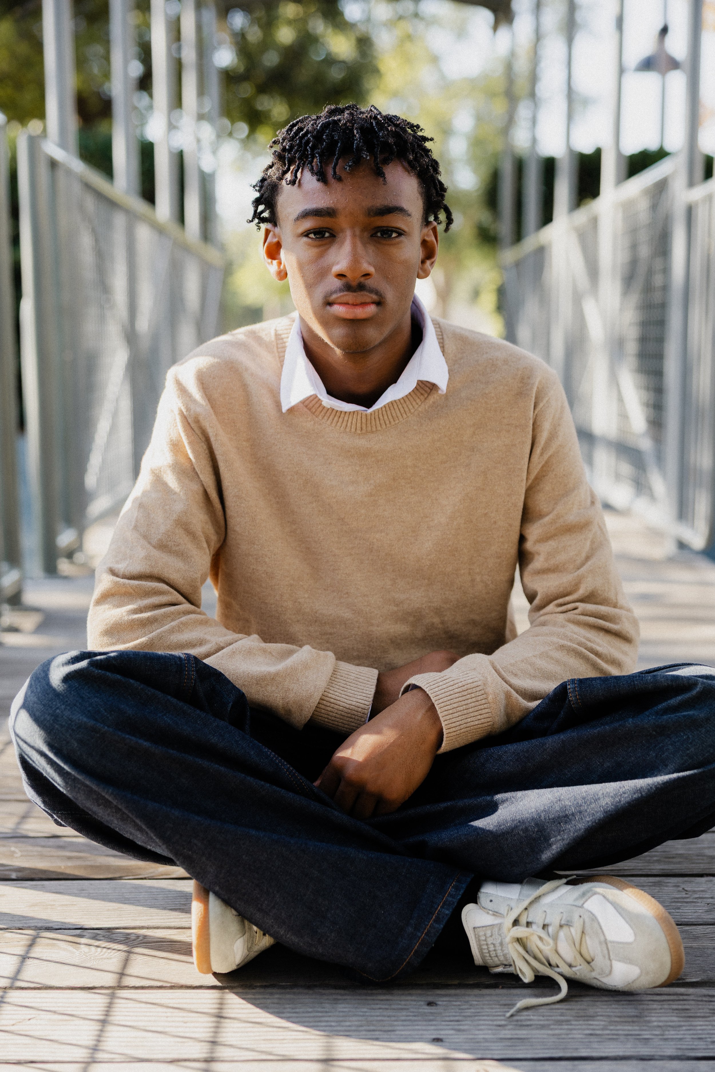 Teenage boy wearing a beige sweater and jeans sitting cross-legged on a wooden walkway at Great Park Irvine.