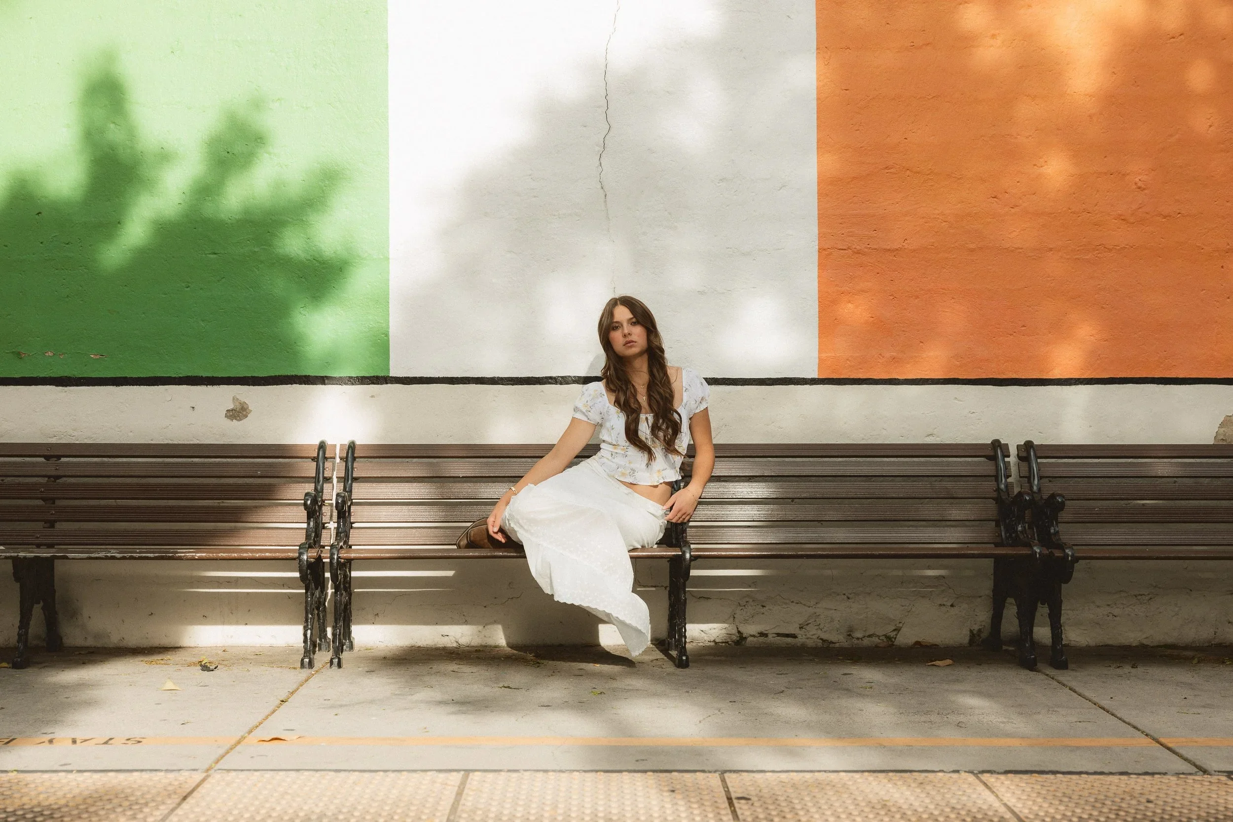A high school senior girl in a floral top and white skirt sitting on a bench in front of a colorful Irish flag wall in the historic Los Rios District of San Juan Capistrano.