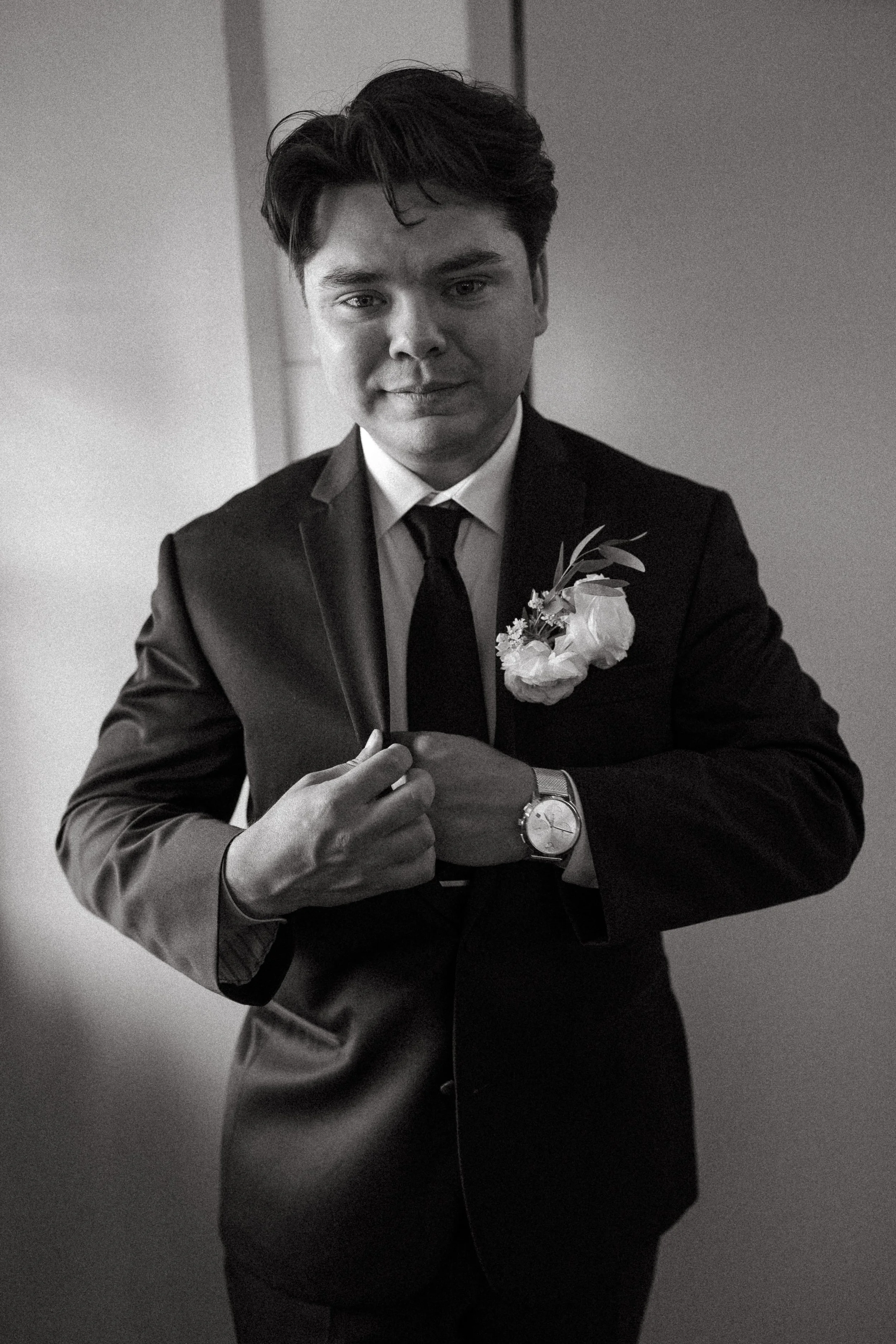 Groom in a black suit and floral boutonniere adjusts his cuff in black and white