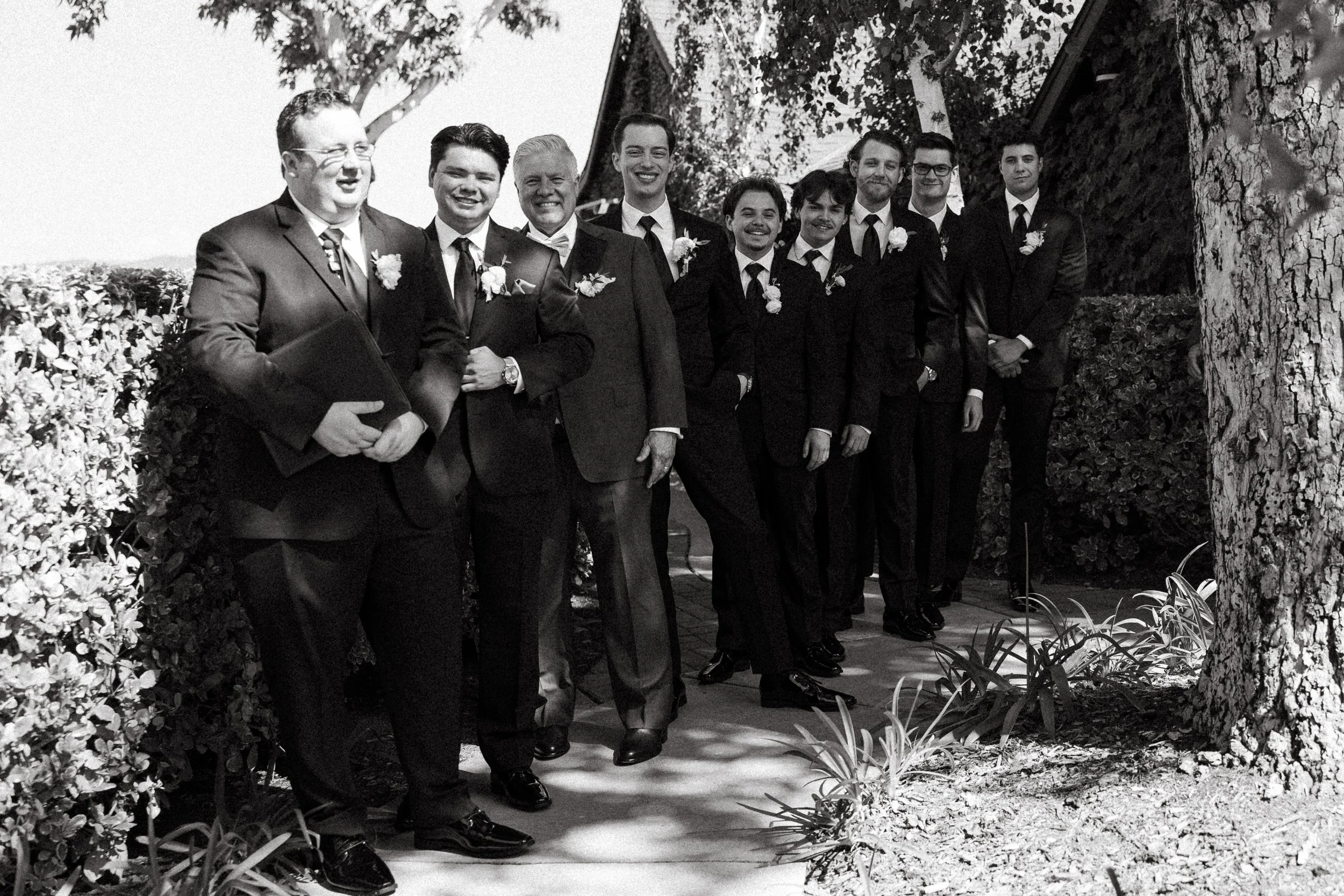 Groom stands with his father, officiant, and groomsmen along a garden path