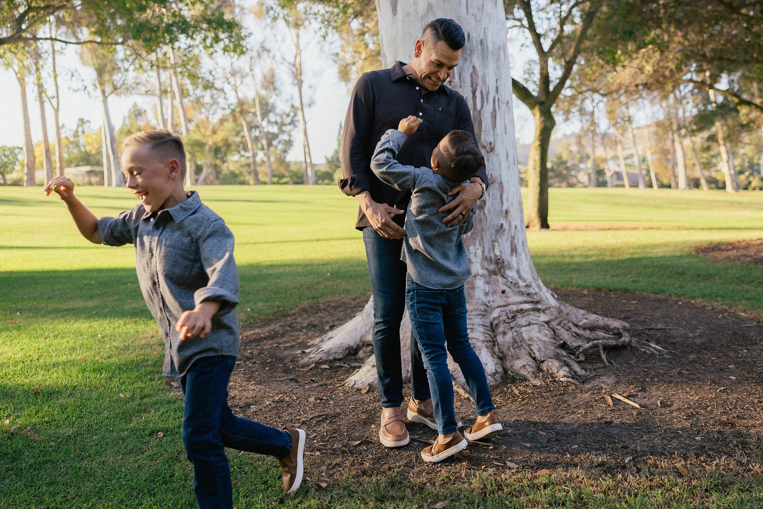 One son runs past while the other hugs his dad near a large tree at Turtle Rock.