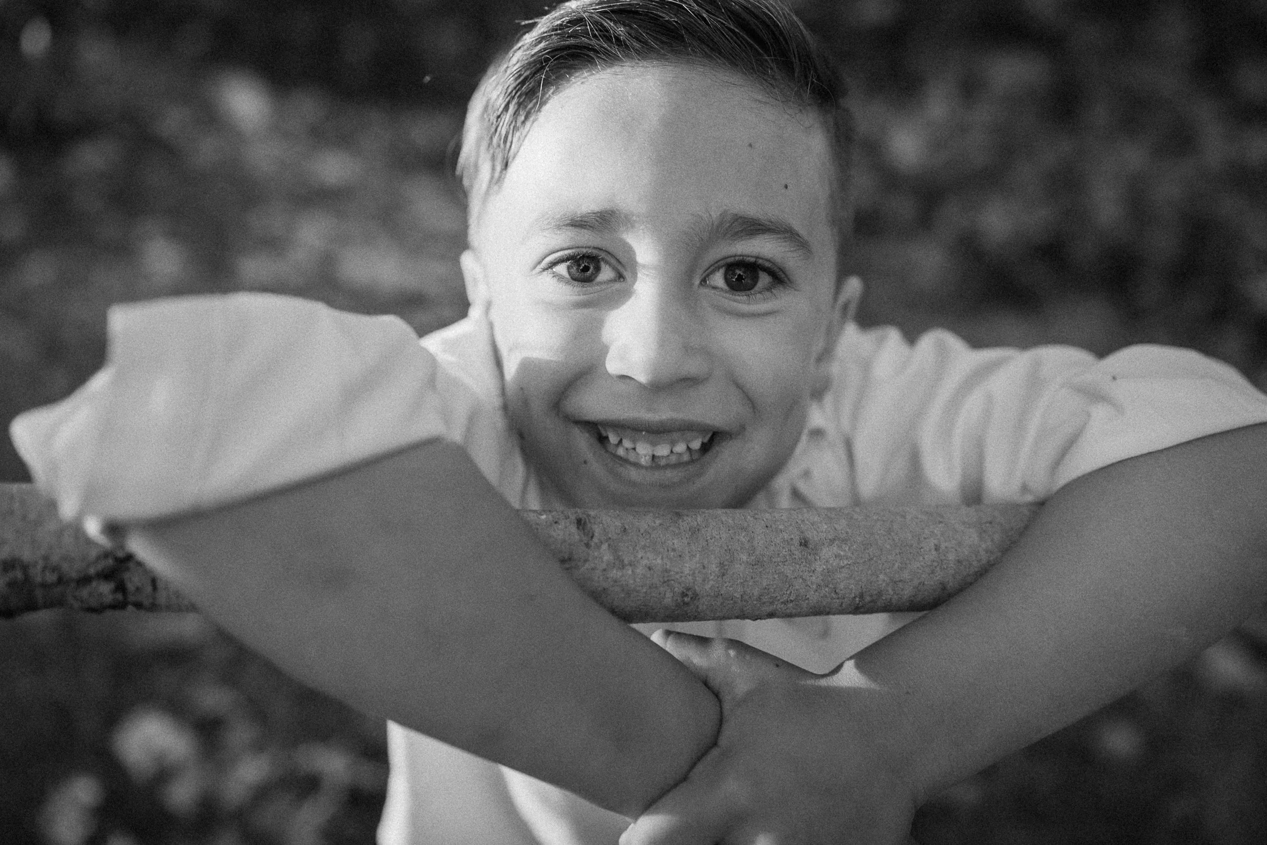 Close-up black-and-white portrait of a smiling boy leaning over a branch.