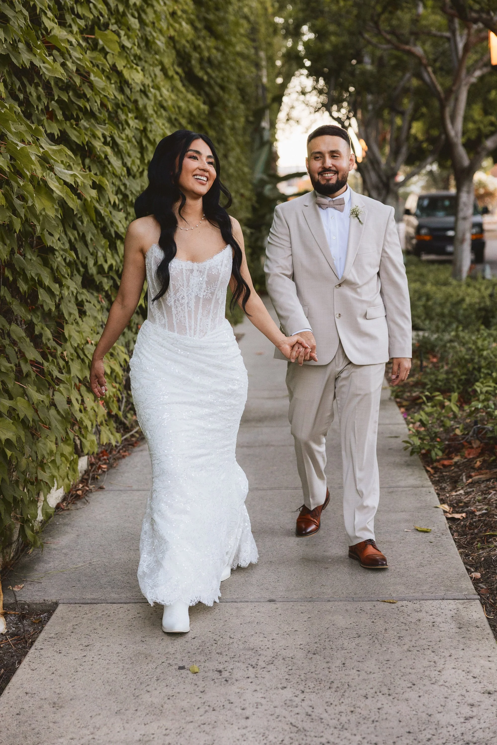Bride and groom walking hand in hand along ivy-covered wall outside Los Angeles River Center and Gardens.