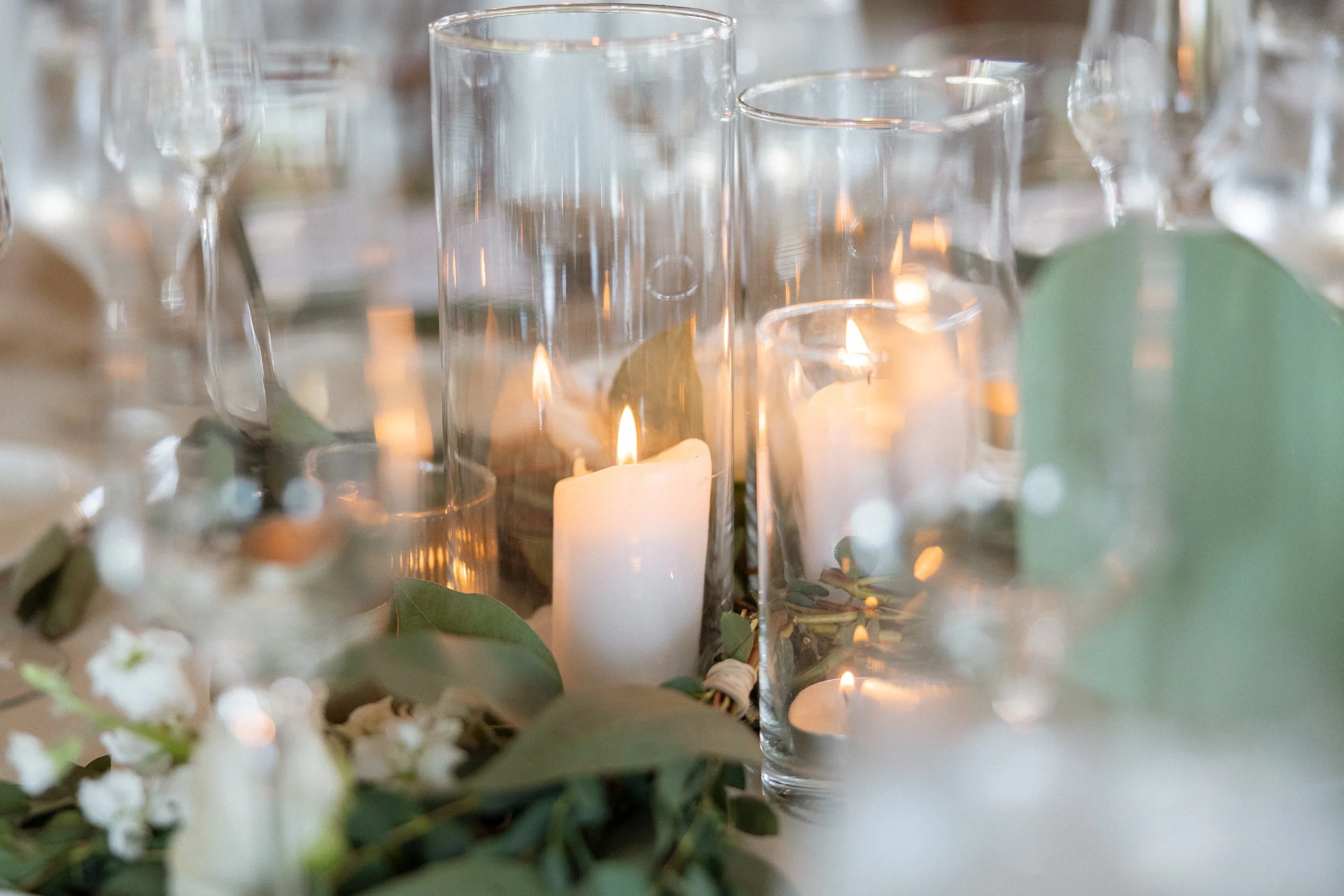 Pillar candles in glass cylinders surrounded by eucalyptus and white blooms