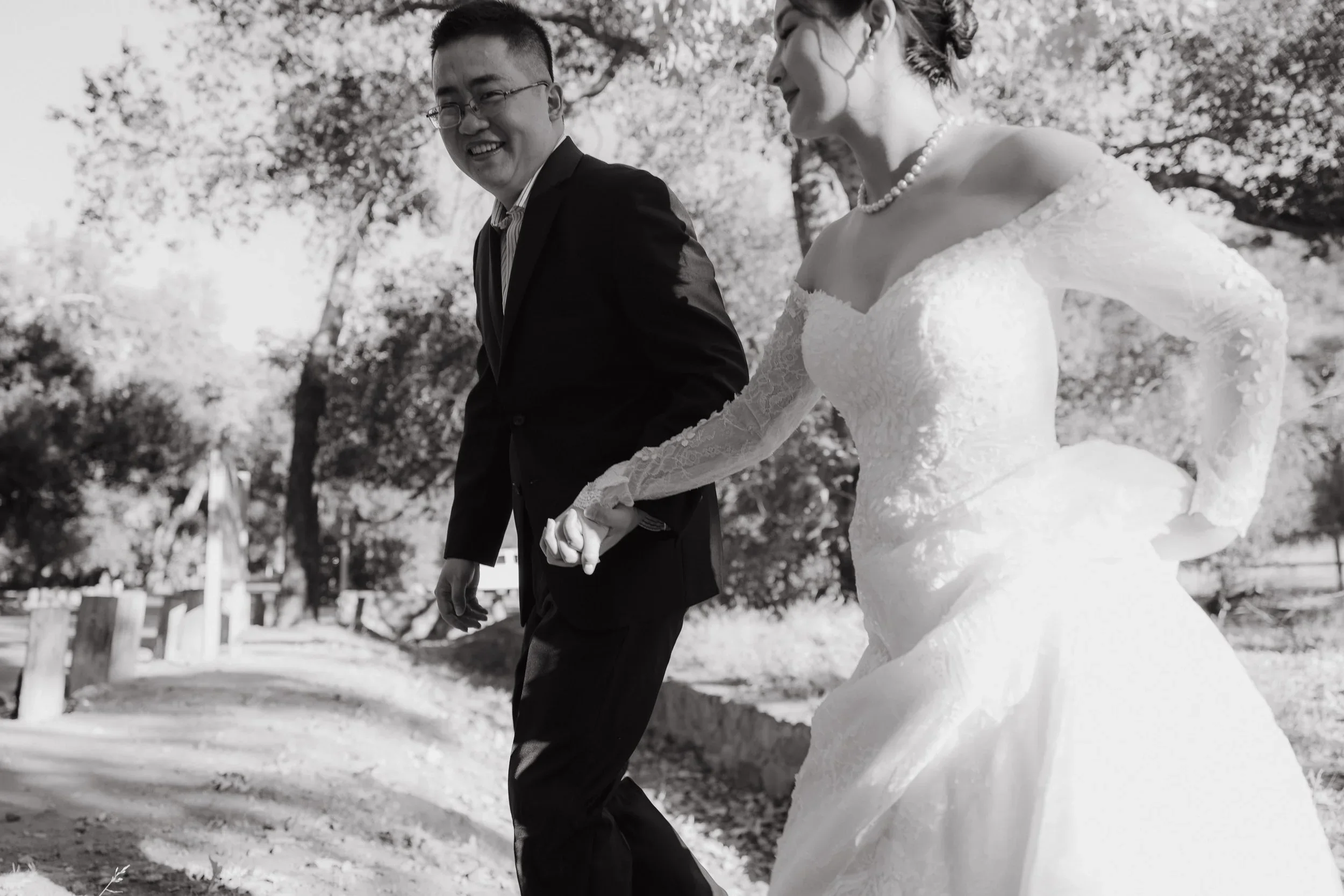 A bride in a white gown and groom in a black suit smiling as they walk along a shaded path at Irvine Regional Park during their wedding portraits.