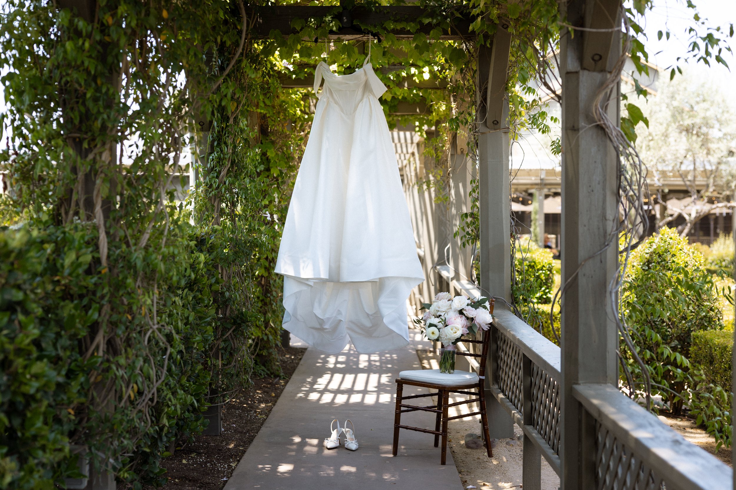 Wedding gown hangs beneath an ivy-covered pergola with shoes and bouquet below