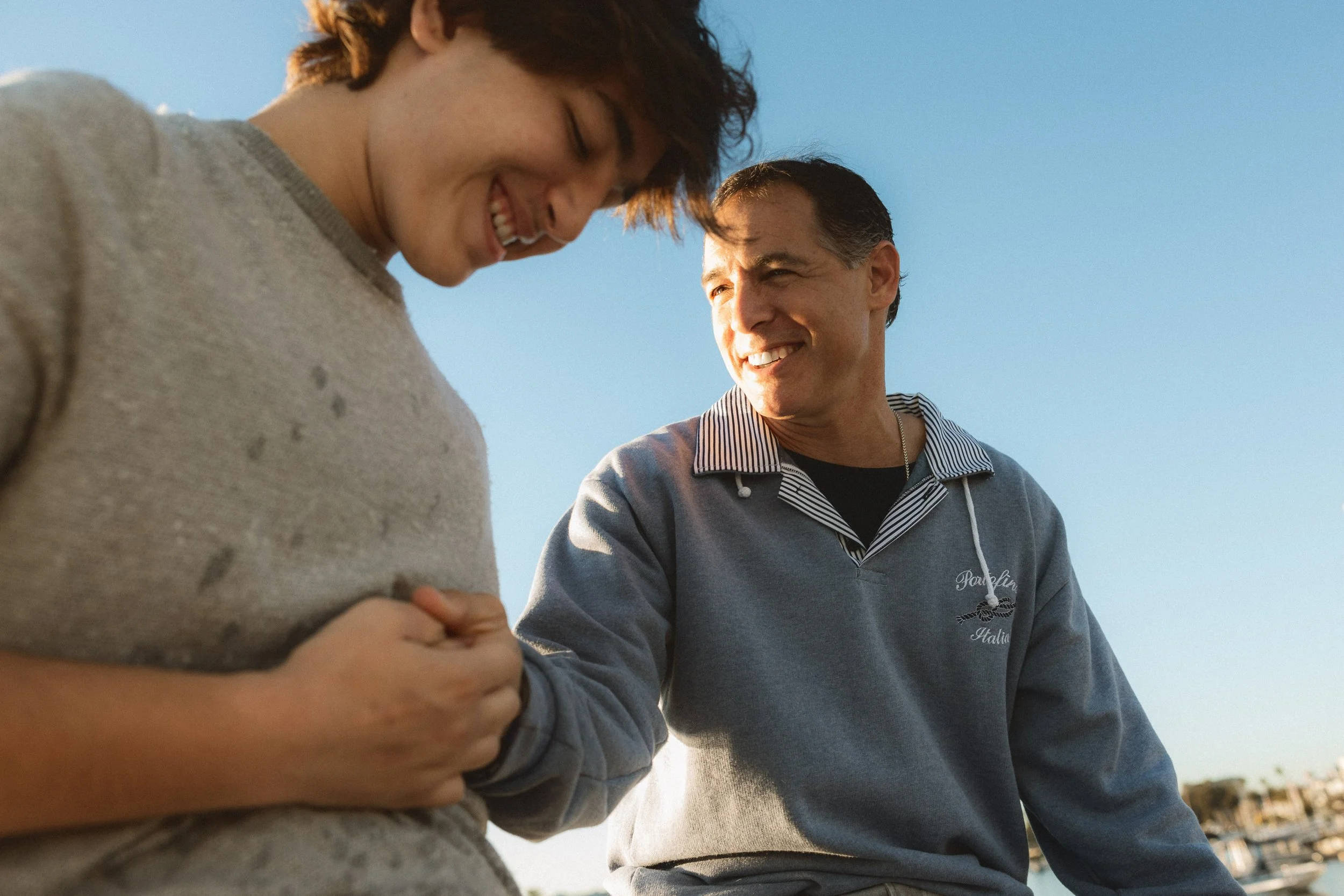 A candid, close-up portrait of a father and his teenage son laughing together on a boat in Newport Harbor during a lifestyle photography session.