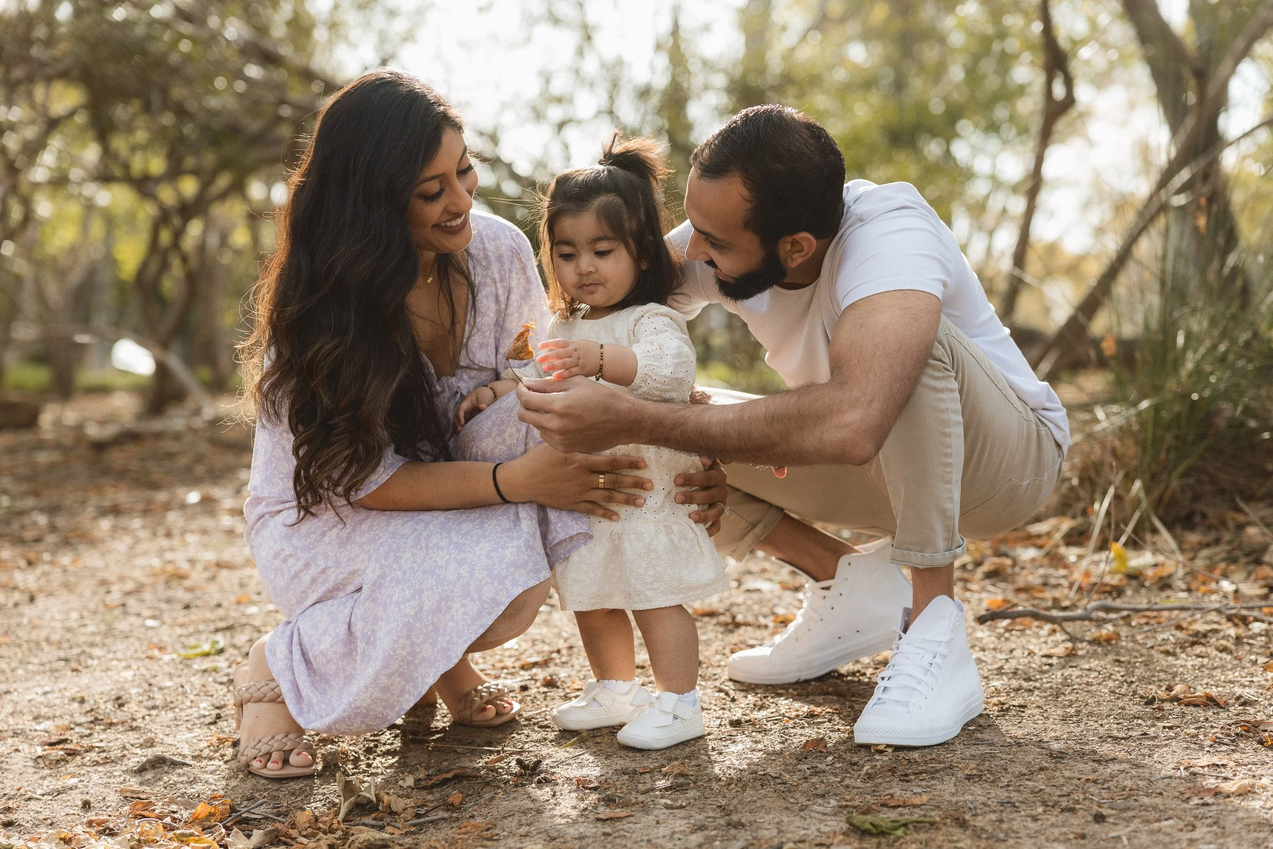 Parents crouching down to hold their toddler daughter's hands on a dirt path under the trees at Jeffrey Open Space Trail.