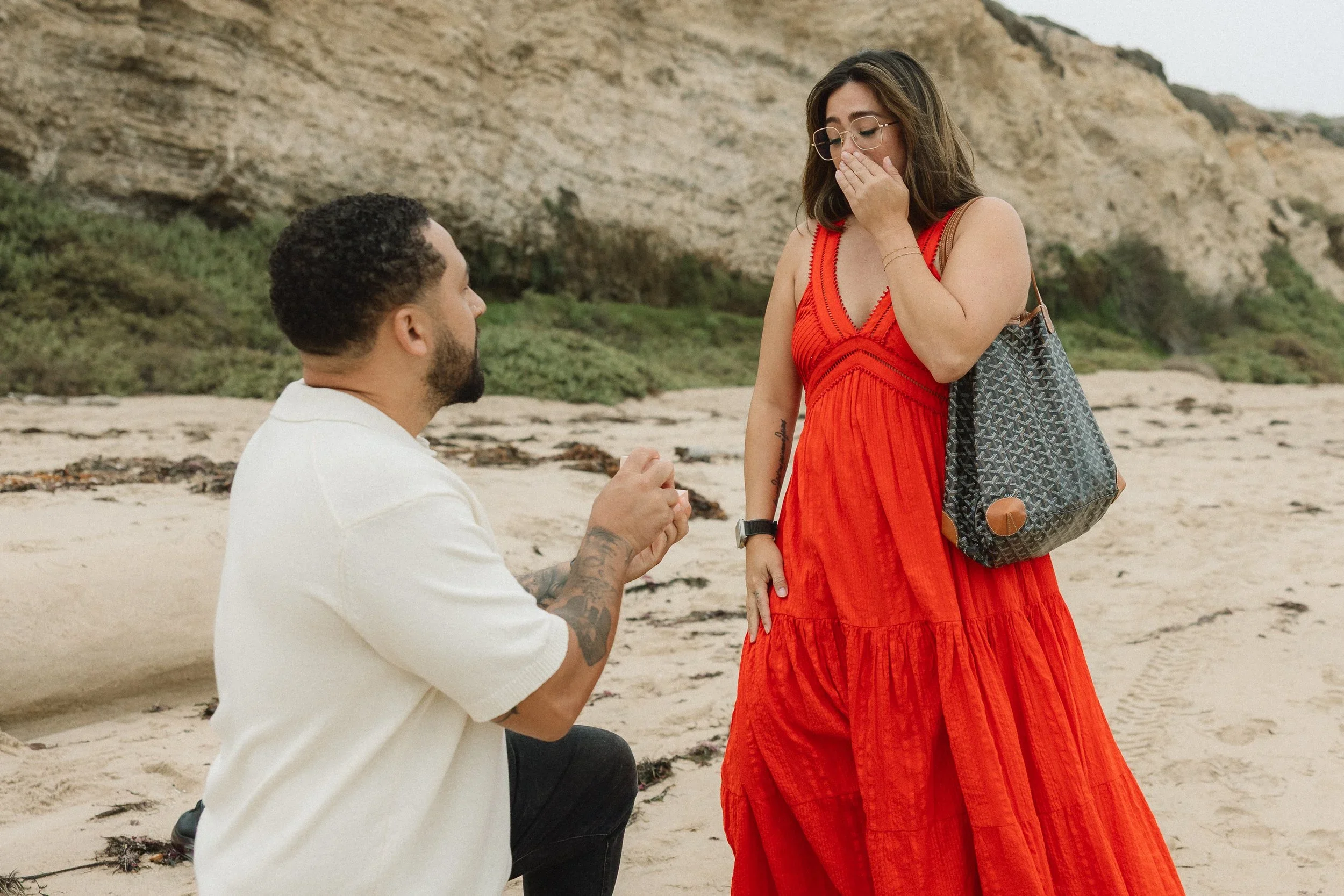 A newly engaged couple on Crystal Cove beach, with the woman reacting in shock and covering her mouth while the man holds the ring box, moments after a surprise proposal.