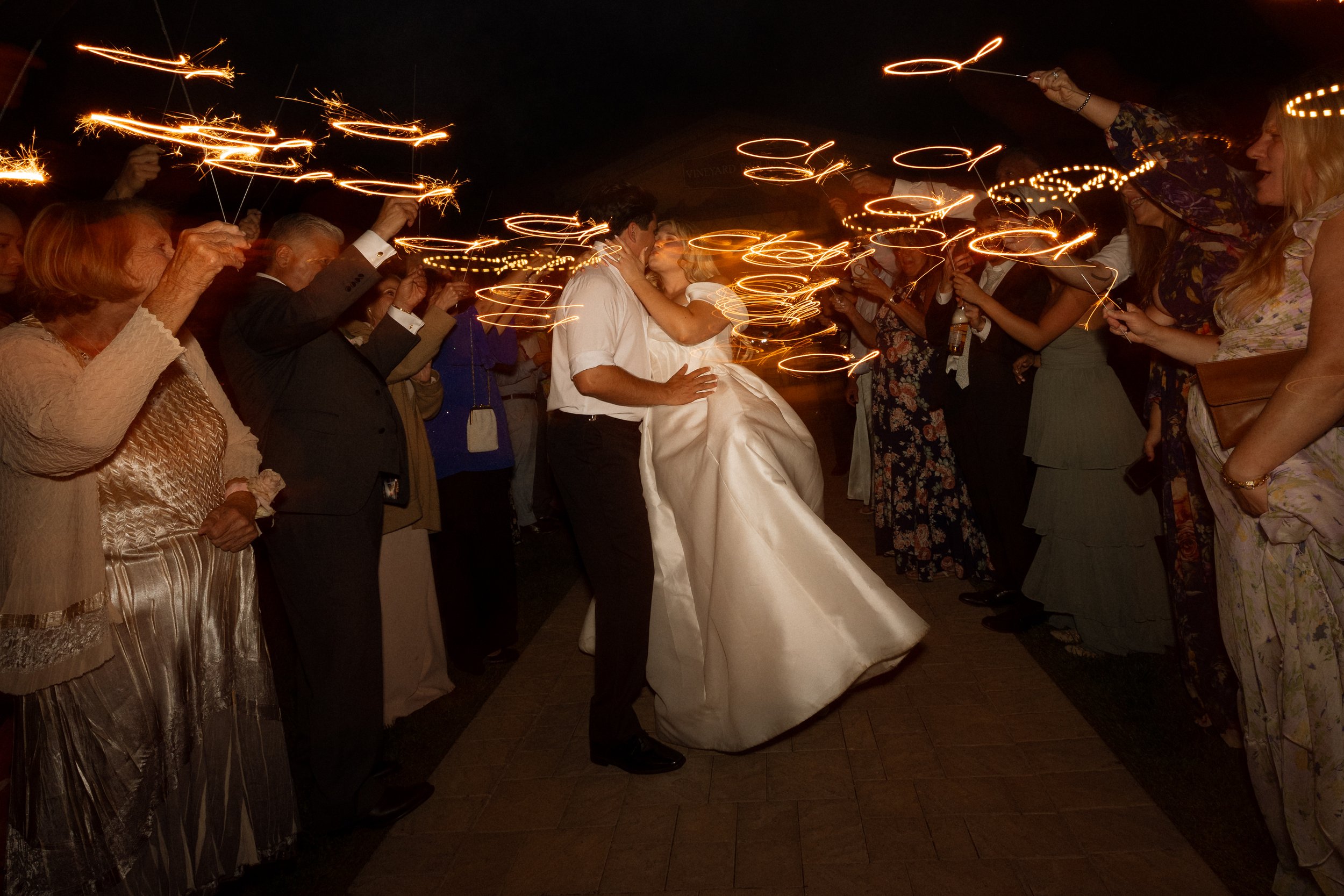 Groom dips the bride for a kiss as guests raise sparklers around them