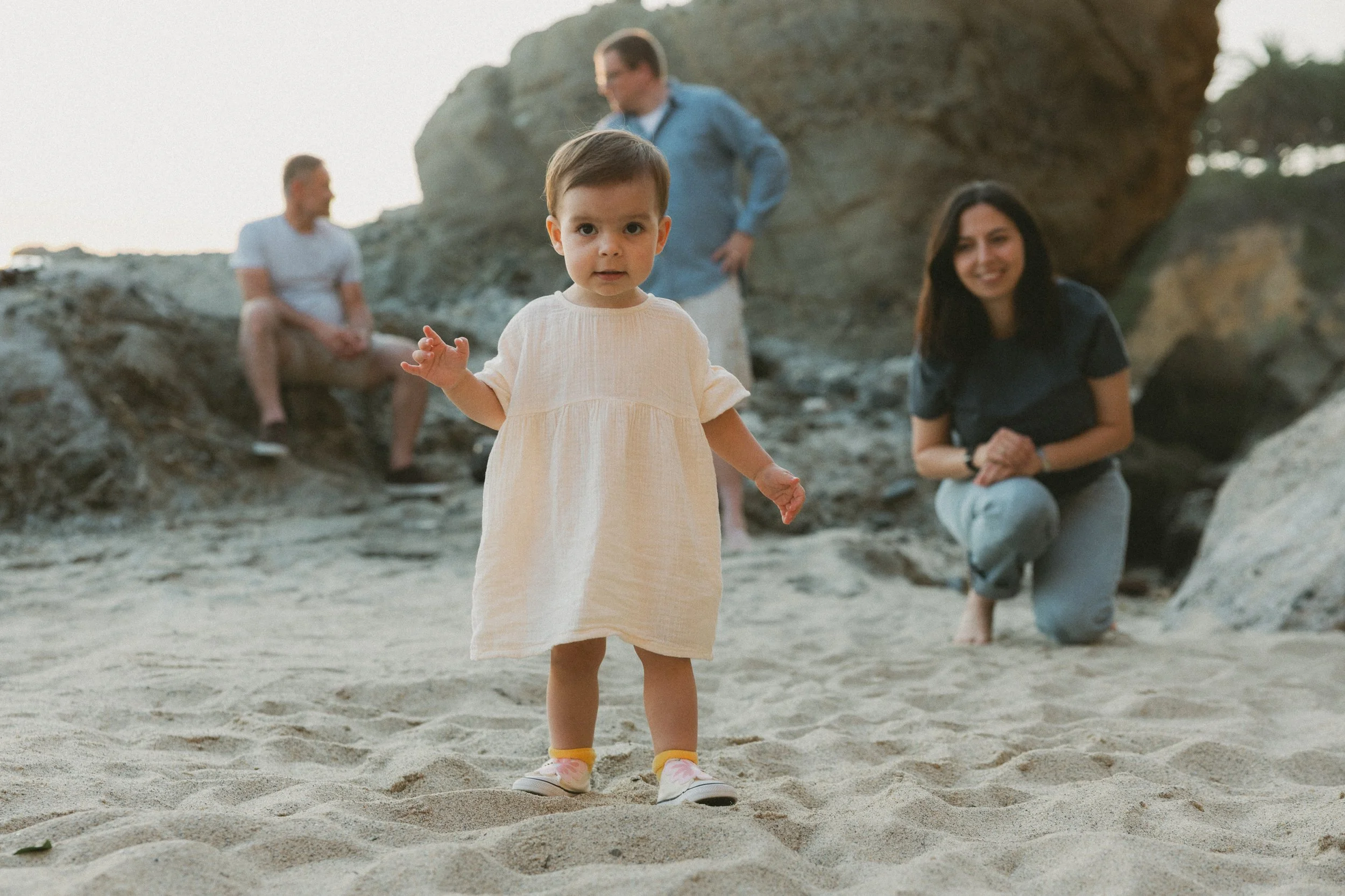 A toddler walking on the sand towards the camera with parents watching in the background during a family session at Treasure Island Beach.