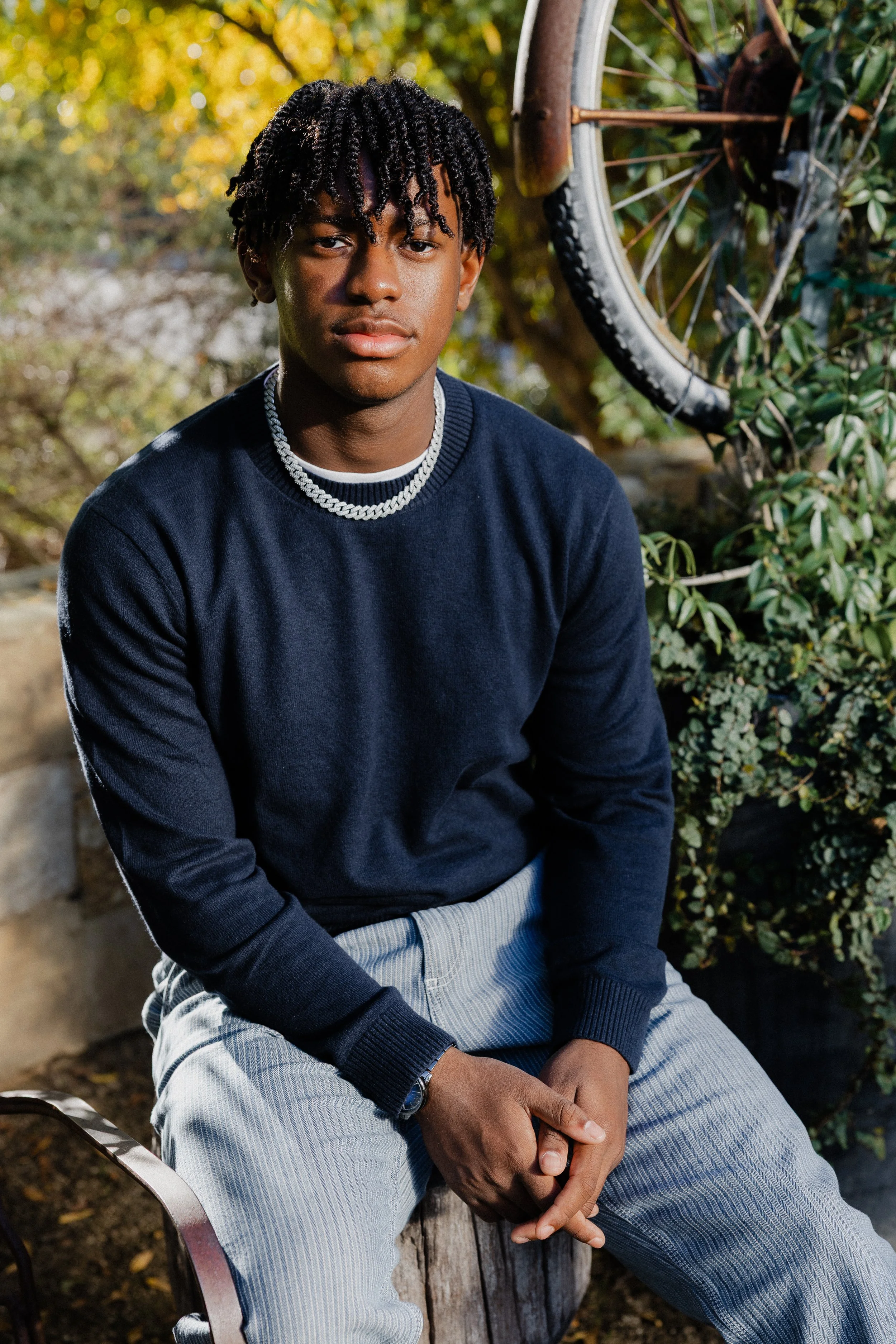 Teenage boy wearing a navy sweater and silver chain sitting on a rustic log at Pavilion Park.