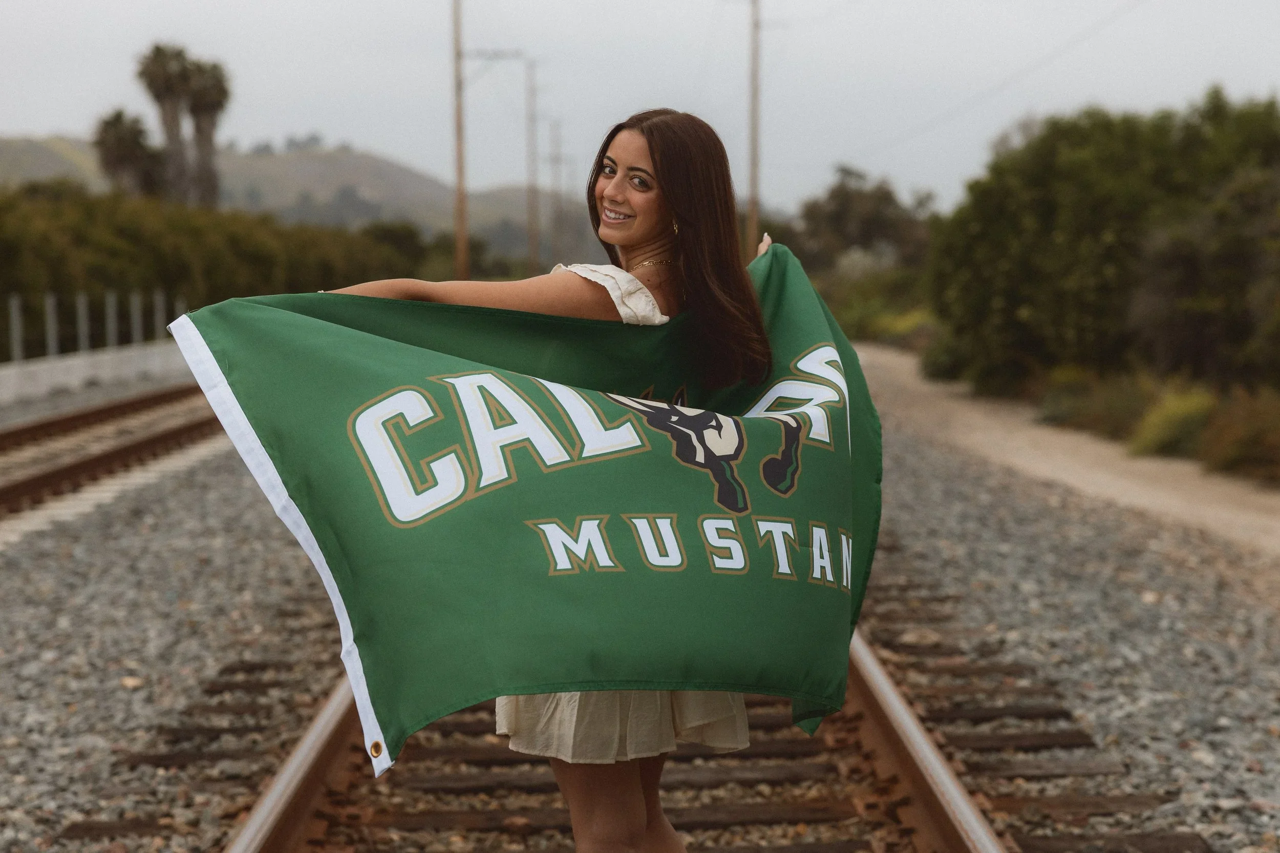 A graduating high school senior girl smiling while holding a green Cal Poly Mustangs flag on train tracks during her college announcement photoshoot.