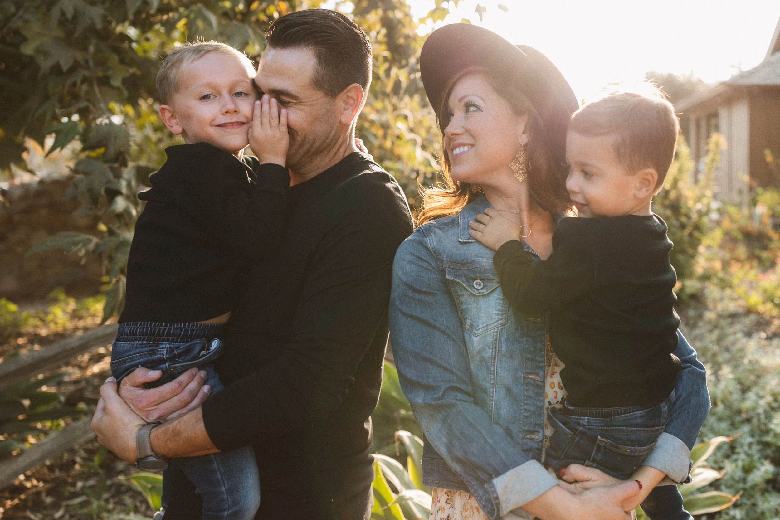 A mother and father holding their two young sons while smiling in the golden afternoon light at the historic San Juan Capistrano Train Station.