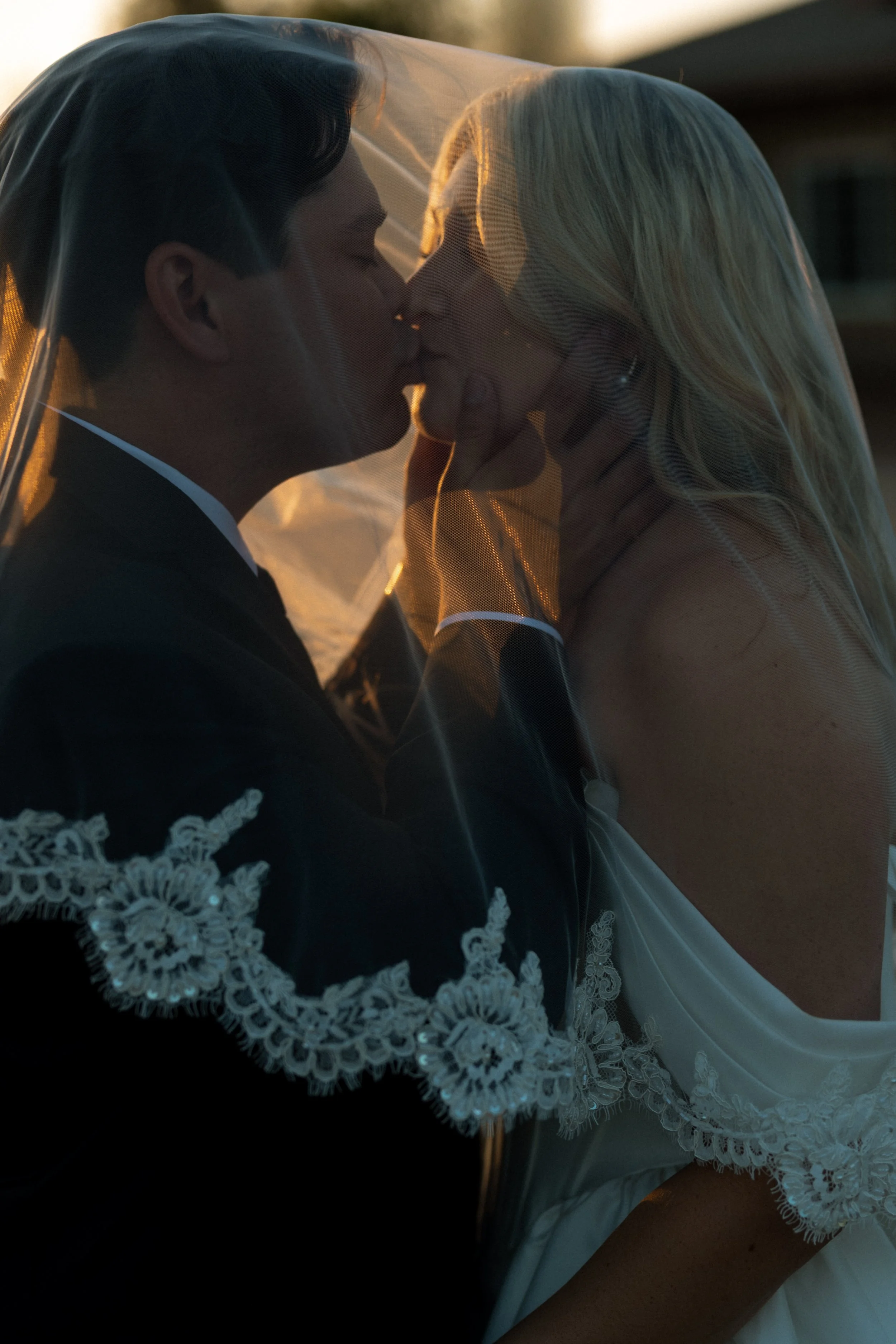 Bride and groom kiss beneath her veil in warm golden hour light