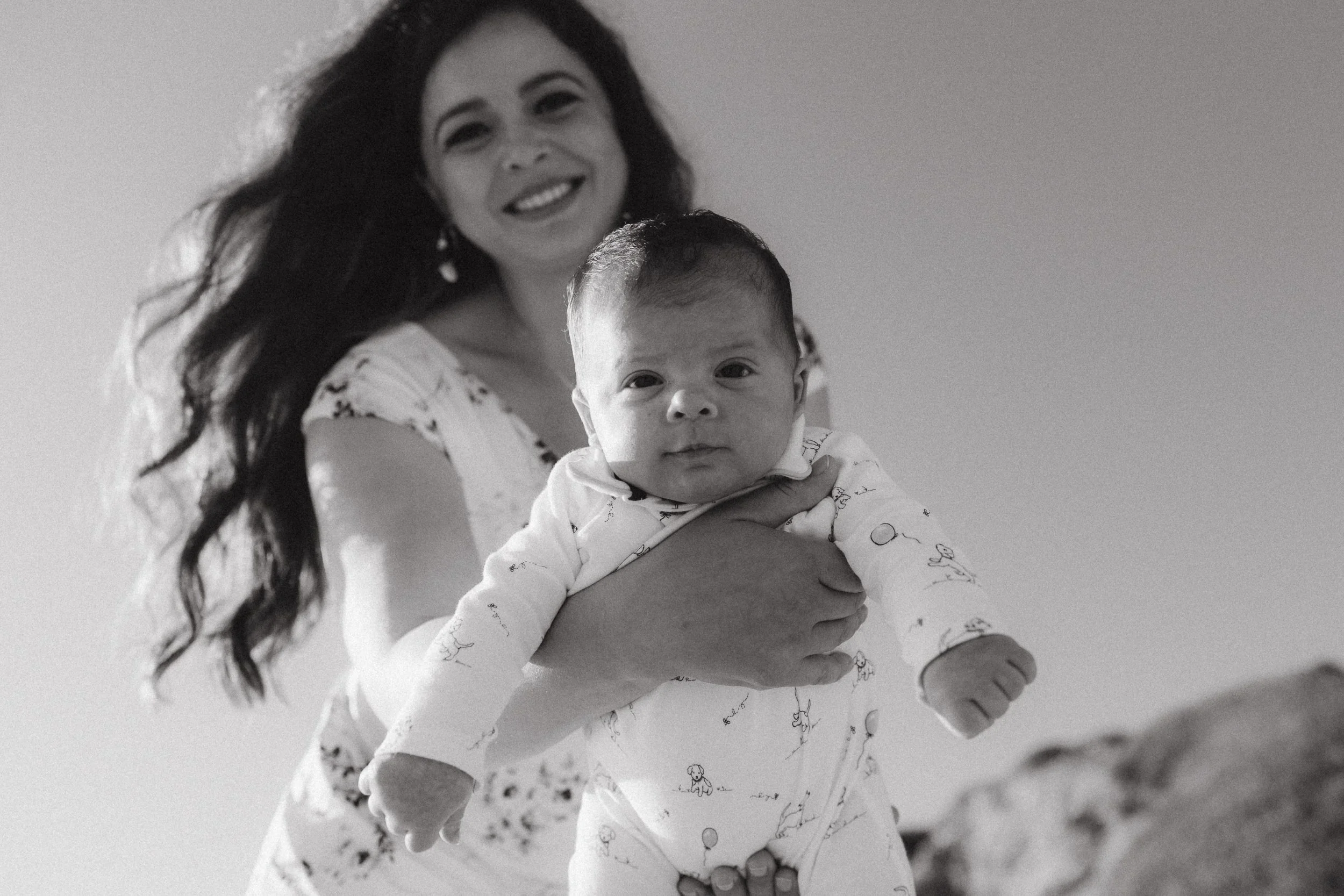 A black and white close-up portrait of a smiling mother holding her baby against the bright sky at Crystal Cove State Park.