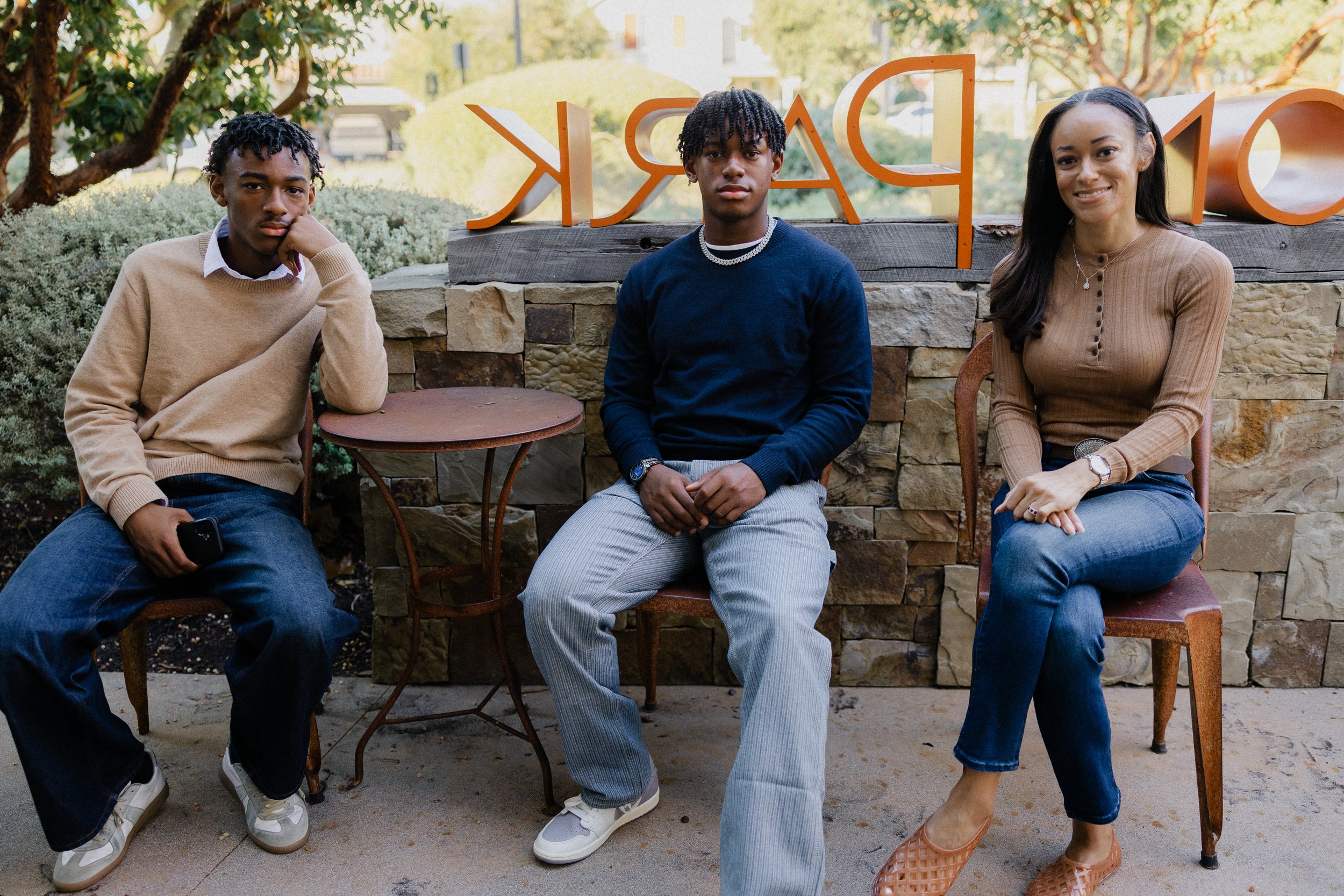 Family of three sitting on rustic chairs in front of the orange "PARK" sign at Pavilion Park in Irvine.