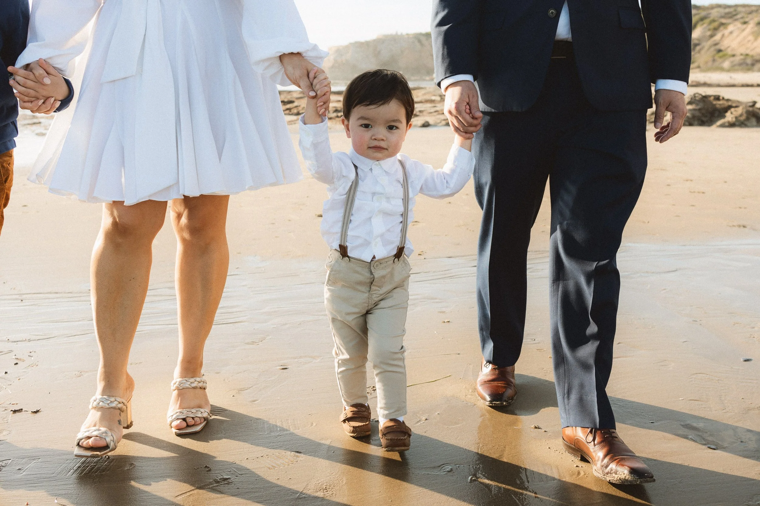 A toddler holding parents' hands while walking on the wet sand during a family session at Crystal Cove State Park.