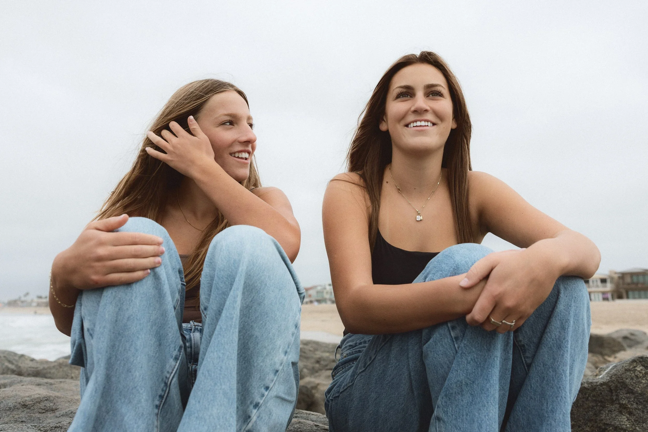 Two teenage sisters sitting on rocks and smiling during a foggy morning family photography session on the Balboa Peninsula in Newport Beach.