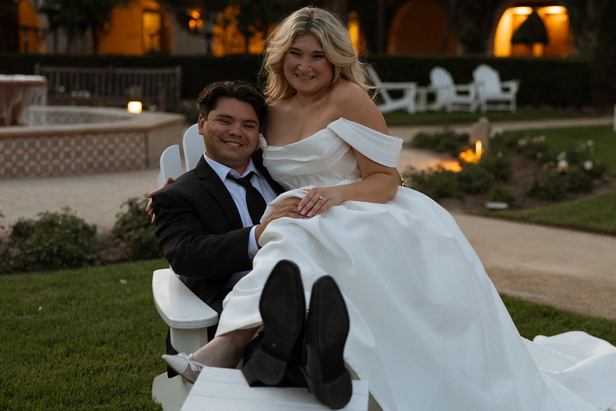 Bride sits across the groom's lap in a garden chair, both laughing