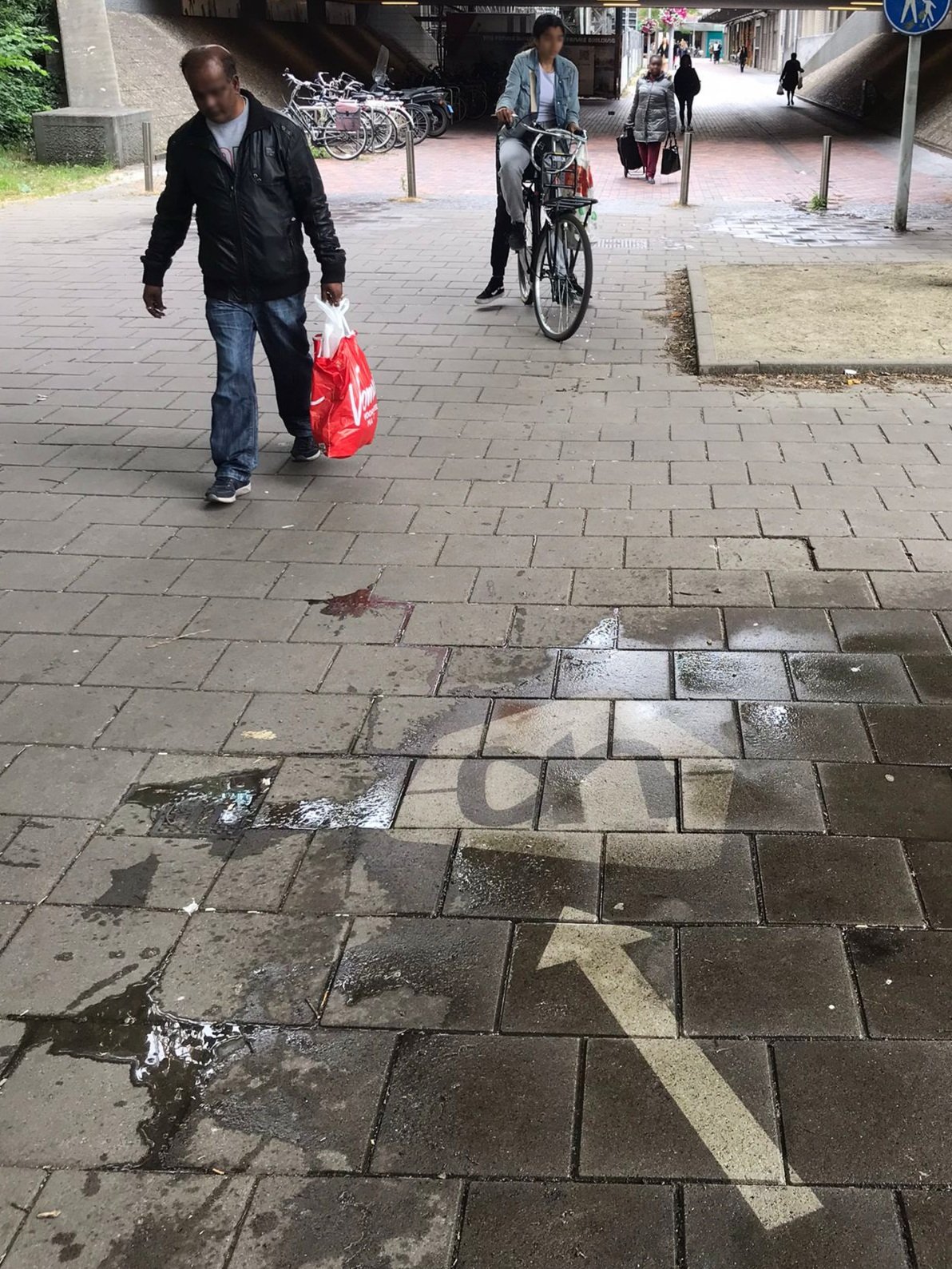 Straat met fietspad en mensen die lopen en fietsen, onder een tunnel met meerdere fietsrekken op de achtergrond. stoepreclame Albert Heijn richting Vomar. In looproute naar concurrent adverteren. op de stoep stoepreclame Greengraffiti .