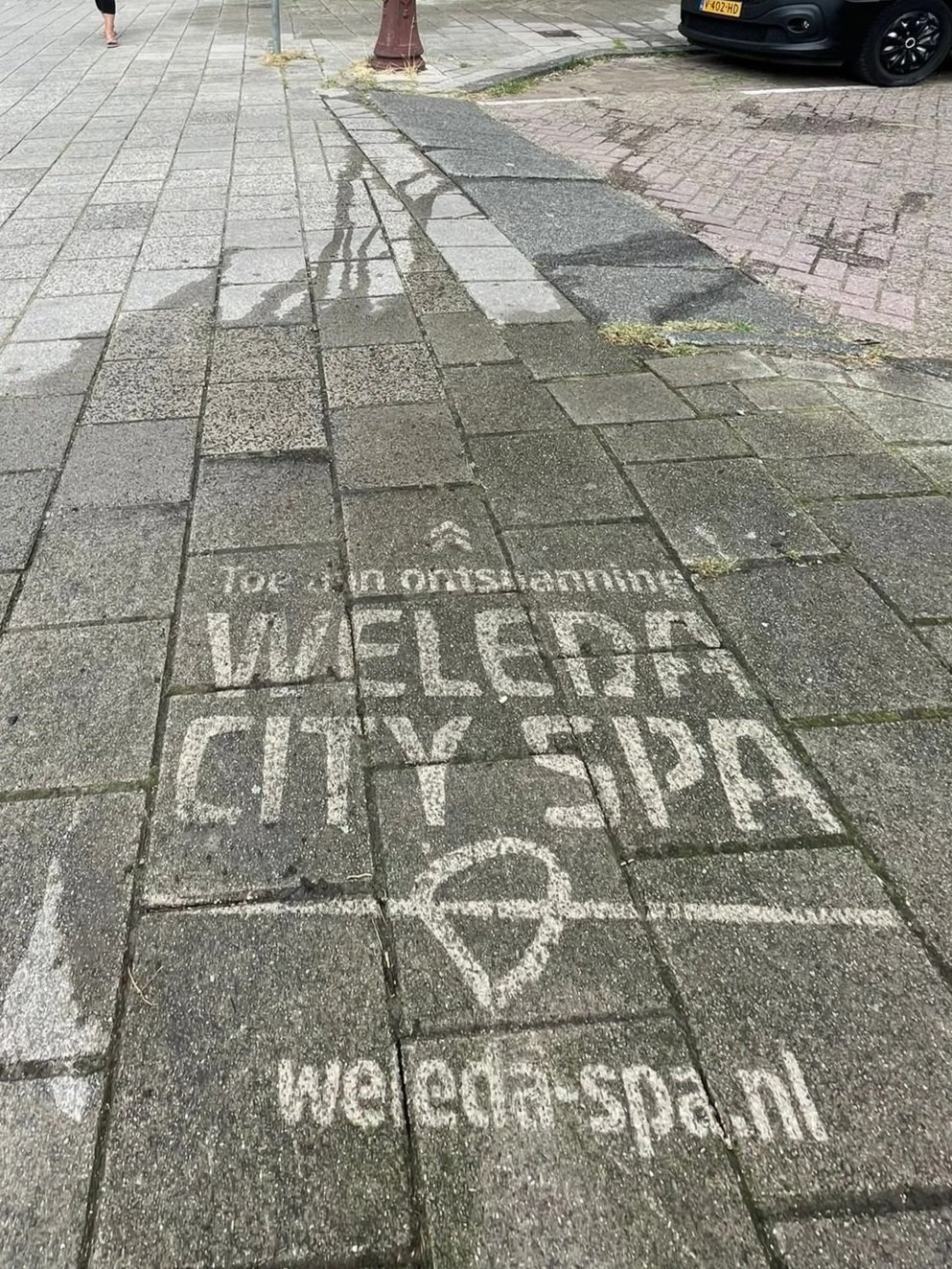 Straat met tegels met witte tekst en een teken, en een website link onderaan. Een voorbeeld van green graffiti ook bekend als clean advertisement, reverse graffiti, stoepreclame van Promendus. straatreclame.