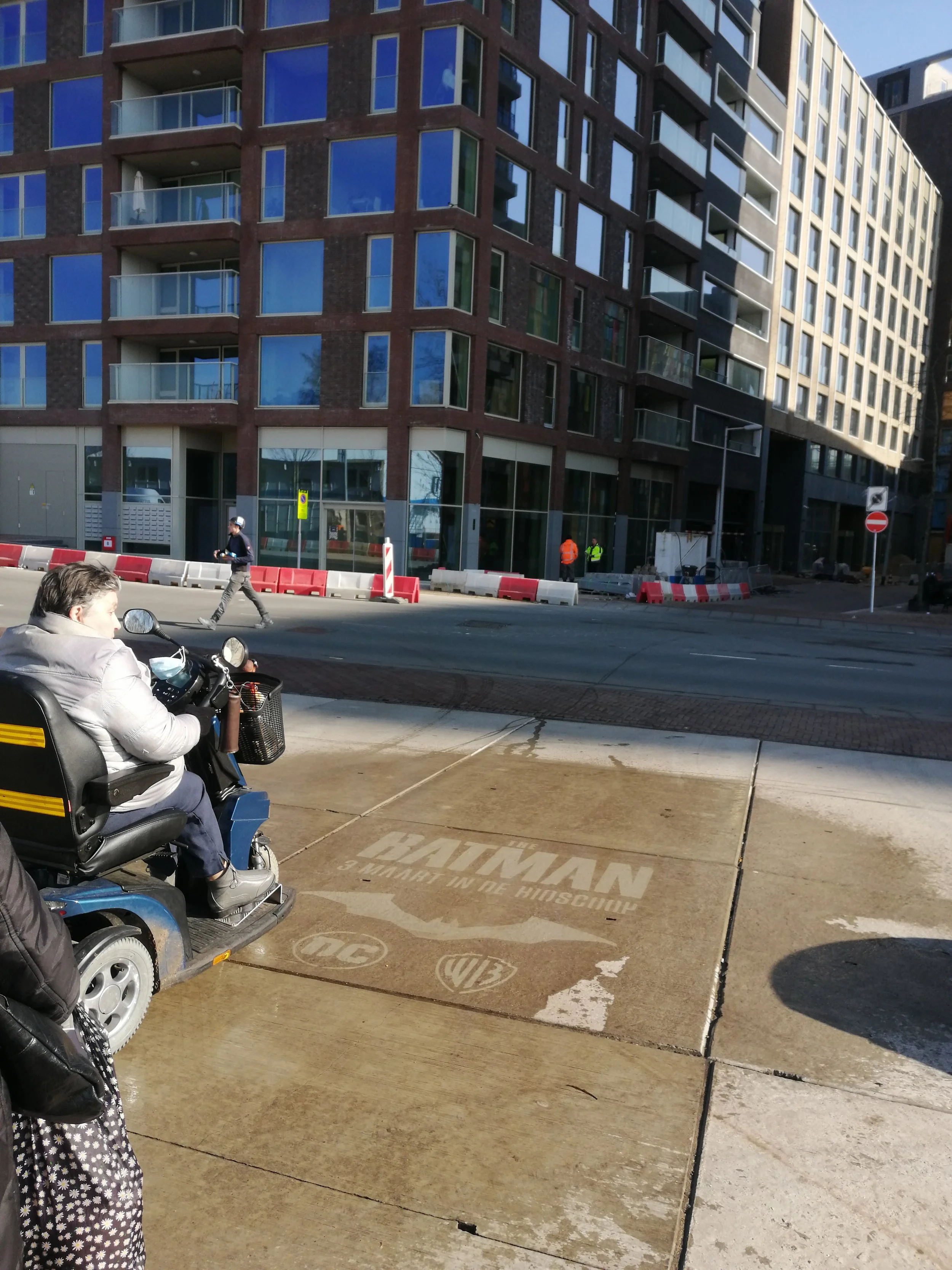 Een straat met een bouwplaats, een vrouw op een scootermobiel en een man die op het trottoir loopt. Op de stoep ligt een logo van Warner Bros. en DC Comics.  clean advertisement, reverse graffiti, stoepreclame van Promendus. guerilla advertising.