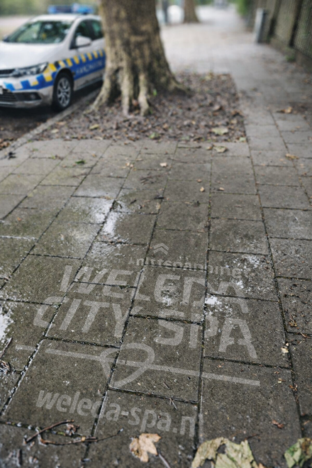 Gele tekst op de stoep die advertentie voor wellness en spa diensten toont, met een politieauto op de achtergrond green graffiti ook bekend als clean advertisement, reverse graffiti, stoepreclame van Promendus.