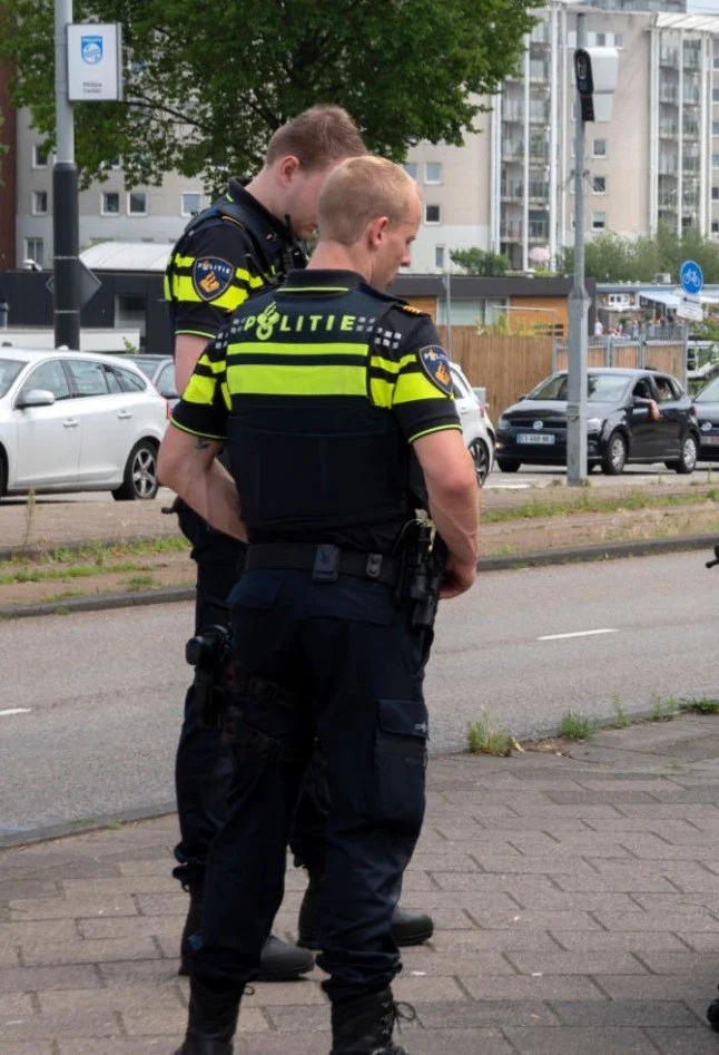 Twee politieagenten staan op een stoep in een stedelijk gebied, dragen uniformen met de tekst 'Politie' en lijken te overleggen of te controleren terwijl ze naar hun telefoons kijken.