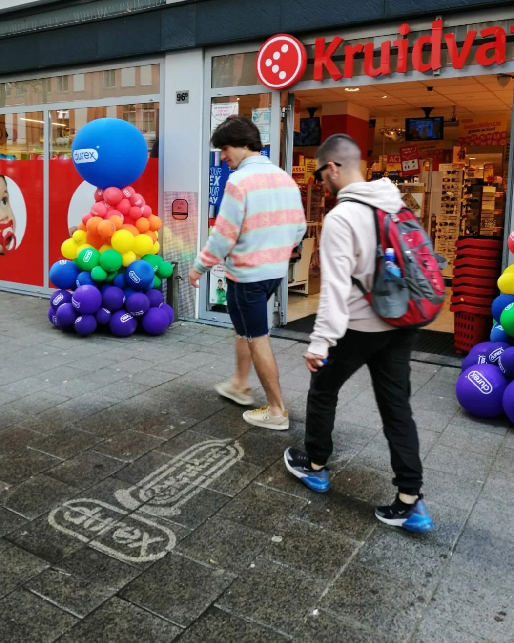 Twee mensen lopen de winkel 'Kruidvat' binnen, met kleurrijke ballonnen buiten de ingang en een 'Durex' banner. Ze dragen sneakers en backpacks. Reclame op de stoep, stoepreclame. Green Graffiti