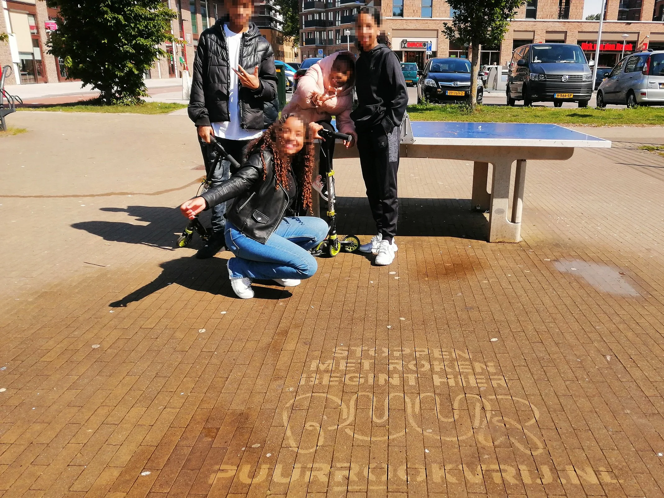 Vijf jongeren poseren onder een pingpongtafel in een park, met een sticker op de grond met teksten.