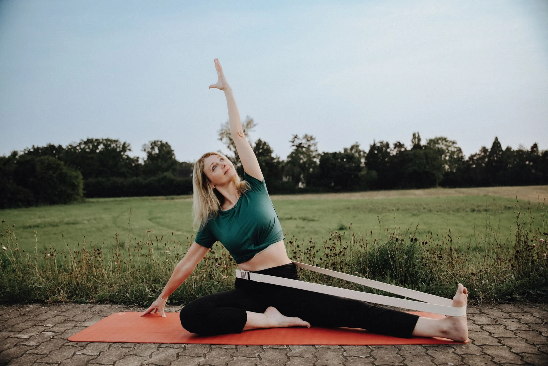 Frau beim Yoga im Freien, dehnt sich mit Widerstandsbanzern auf einer Yogamatte auf einem gepflasterten Weg, im Hintergrund eine Wiese und Bäume, bewölkter Himmel.