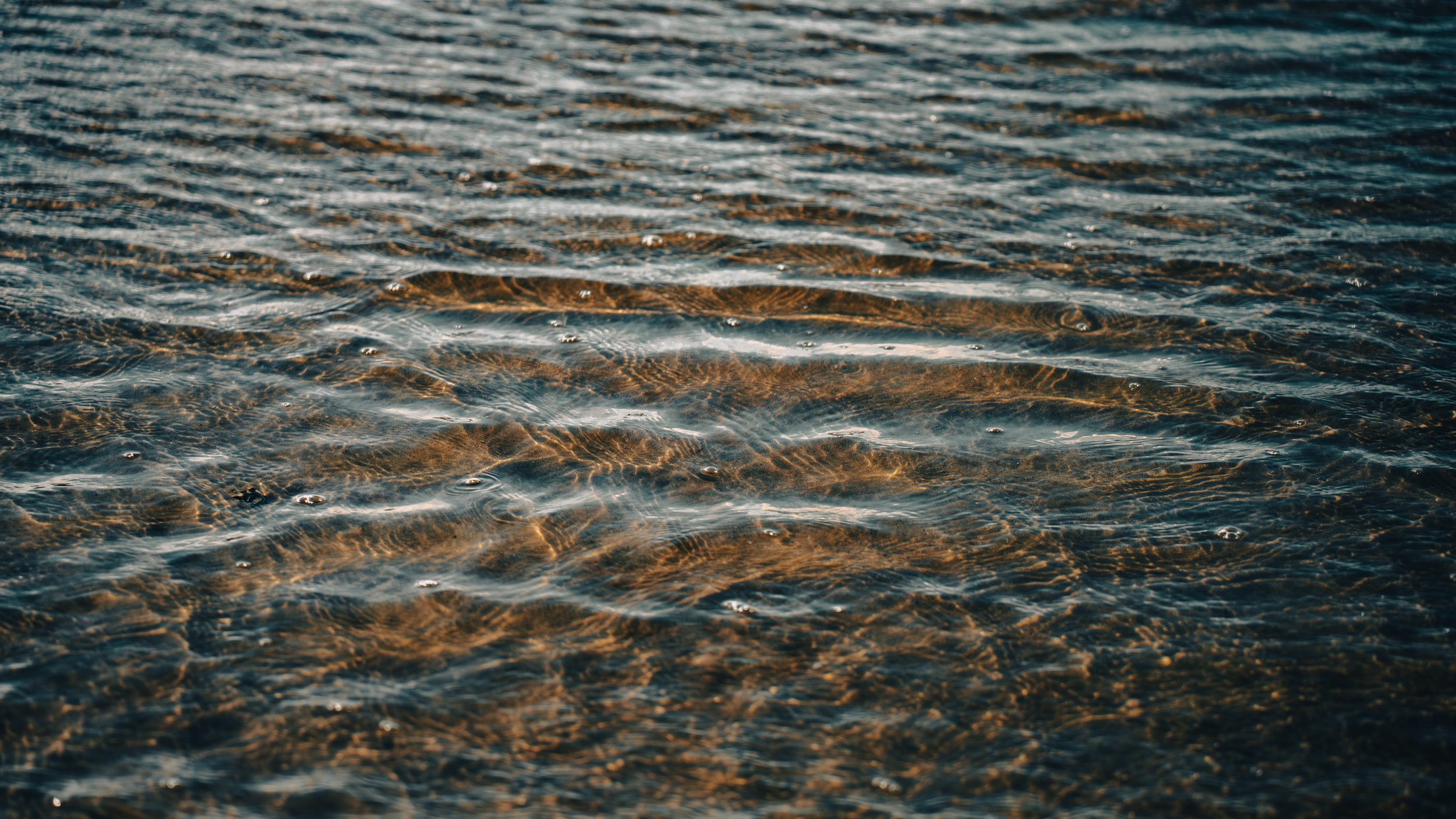 Close-up of gently rippling water with sunlight reflections, showing sand beneath.