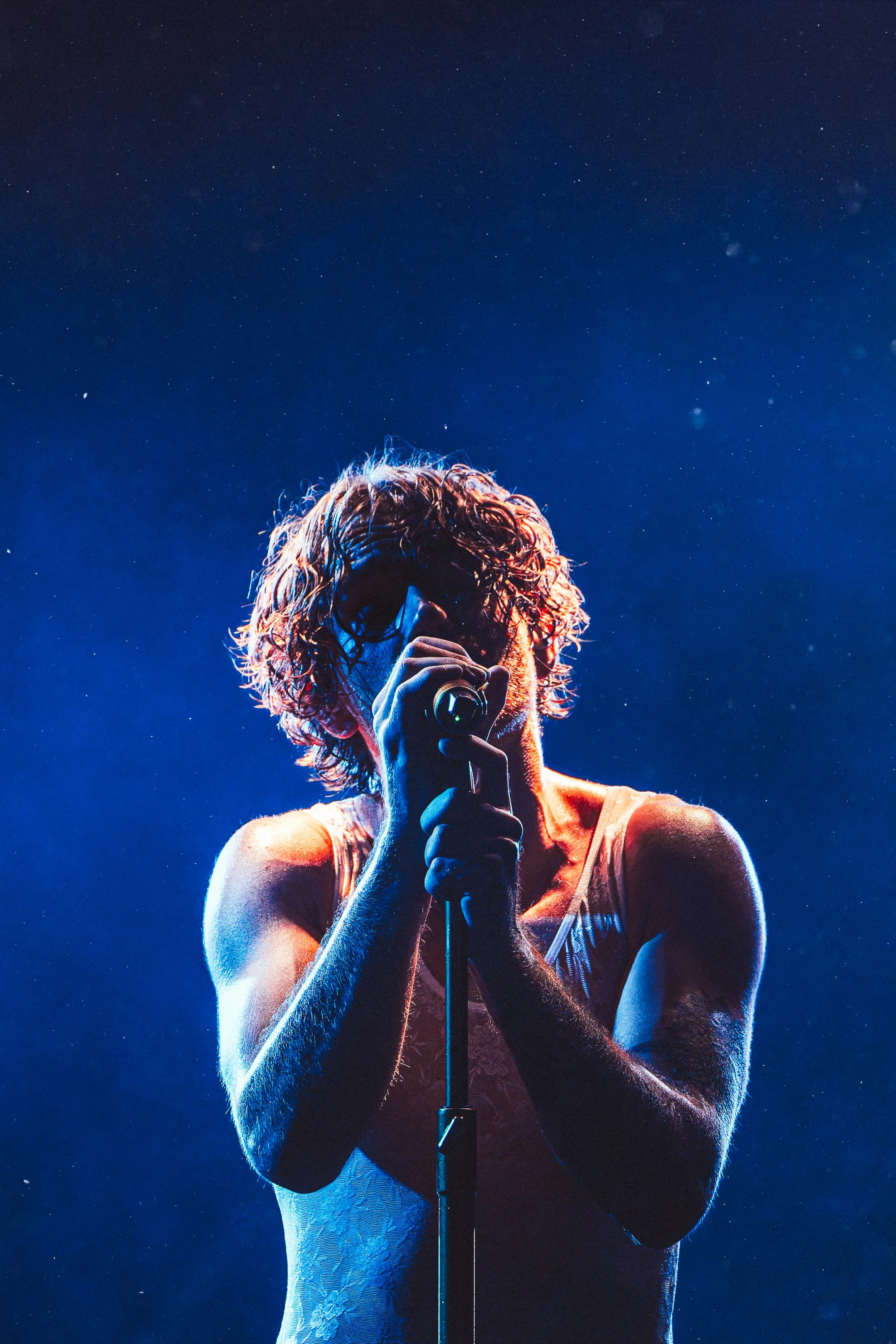 A male singer with curly hair performing on stage, illuminated by blue stage lights, holding a microphone close to his mouth.