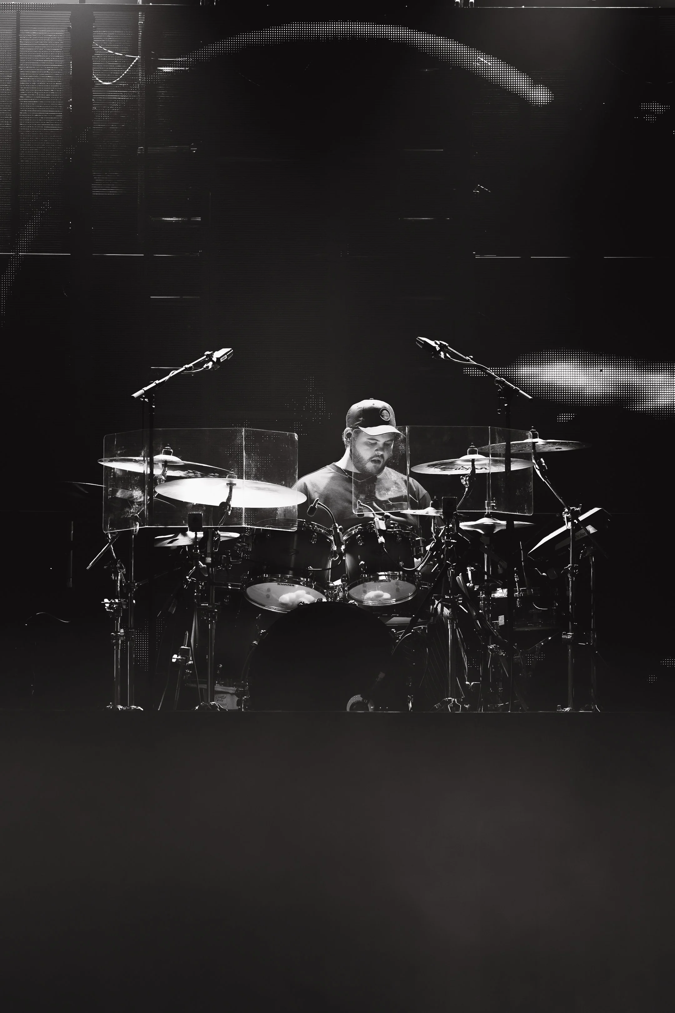 A musician wearing a baseball cap plays a drum set on a stage, with microphones and transparent drum shields around the drums, illuminated by stage lights in a dark setting.
