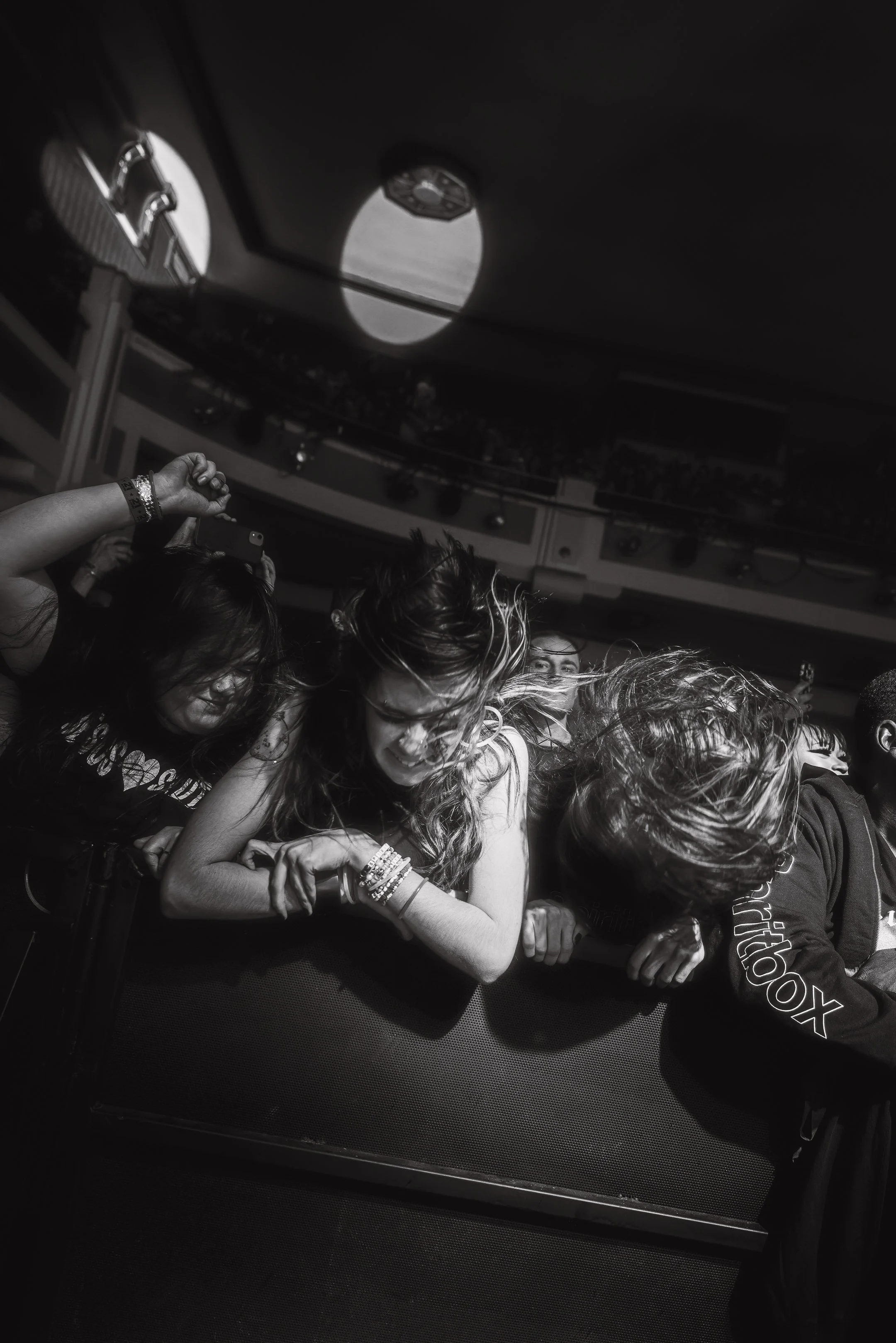 Black and white photo of people at a concert, visibly enjoying and dancing near the stage with hair moving, and some holding phones to capture the moment.