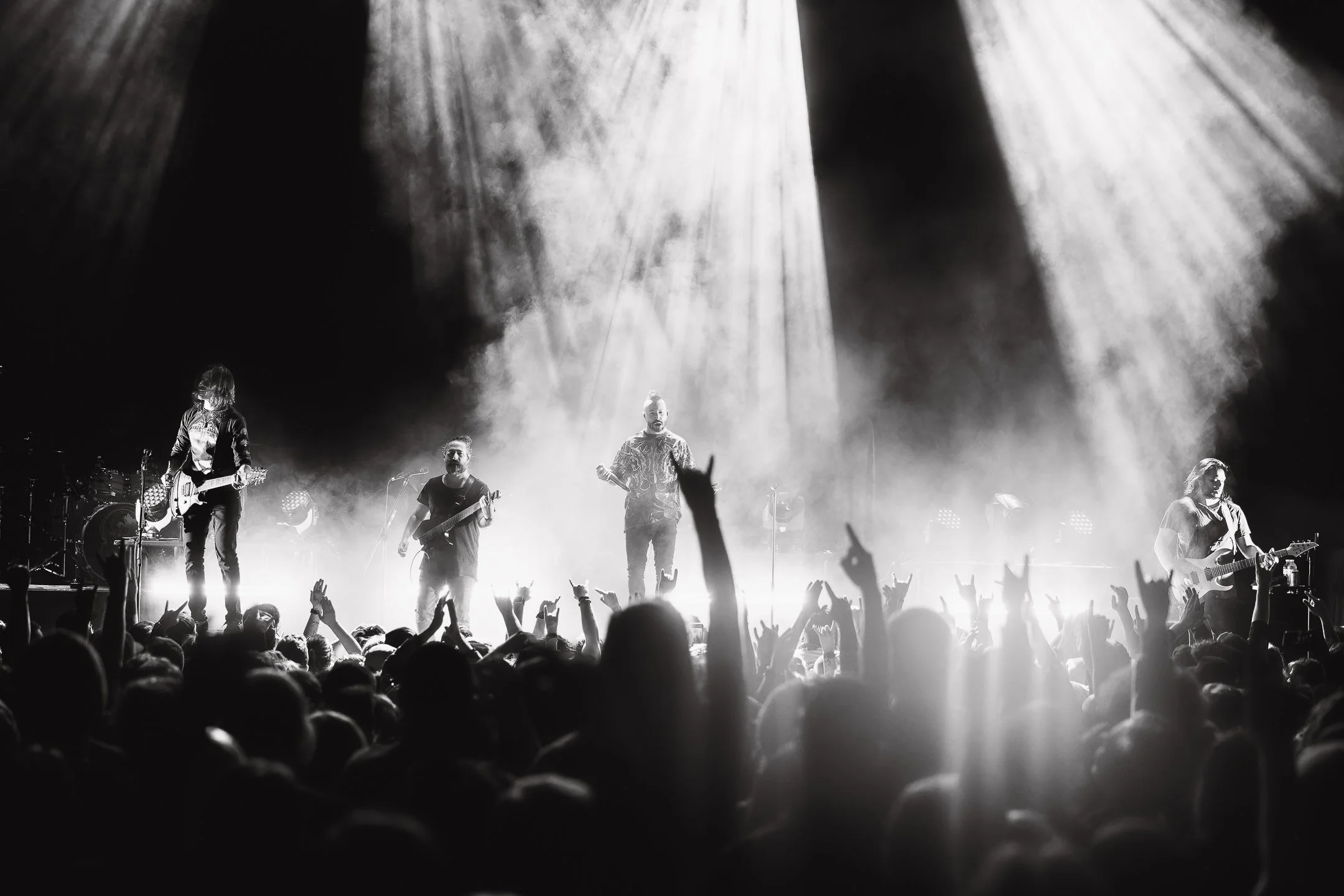 Black and white photo of a live concert with a band performing on stage, surrounded by an enthusiastic crowd with raised hands.