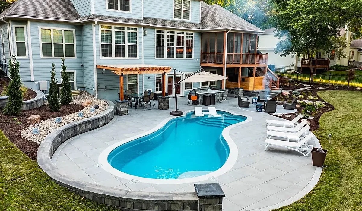 Backyard with a kidney-shaped swimming pool surrounded by loungers and patio furniture; a pergola with a dining set is attached to a two-story house with a screened porch. Landscaping includes neatly trimmed grass and shrubs.