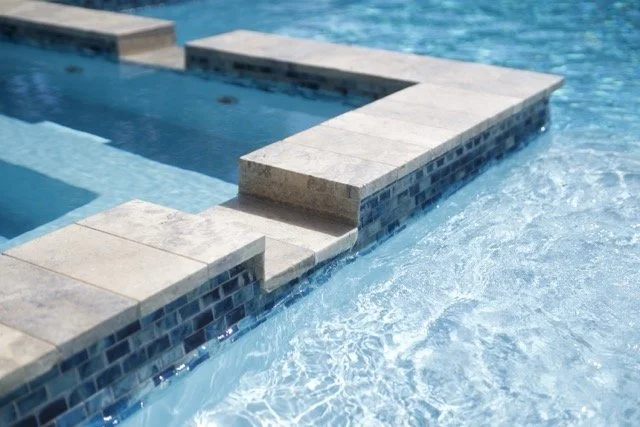 Close-up of a swimming pool, showing a stone-tiled edge and clear blue water in a stepped design.