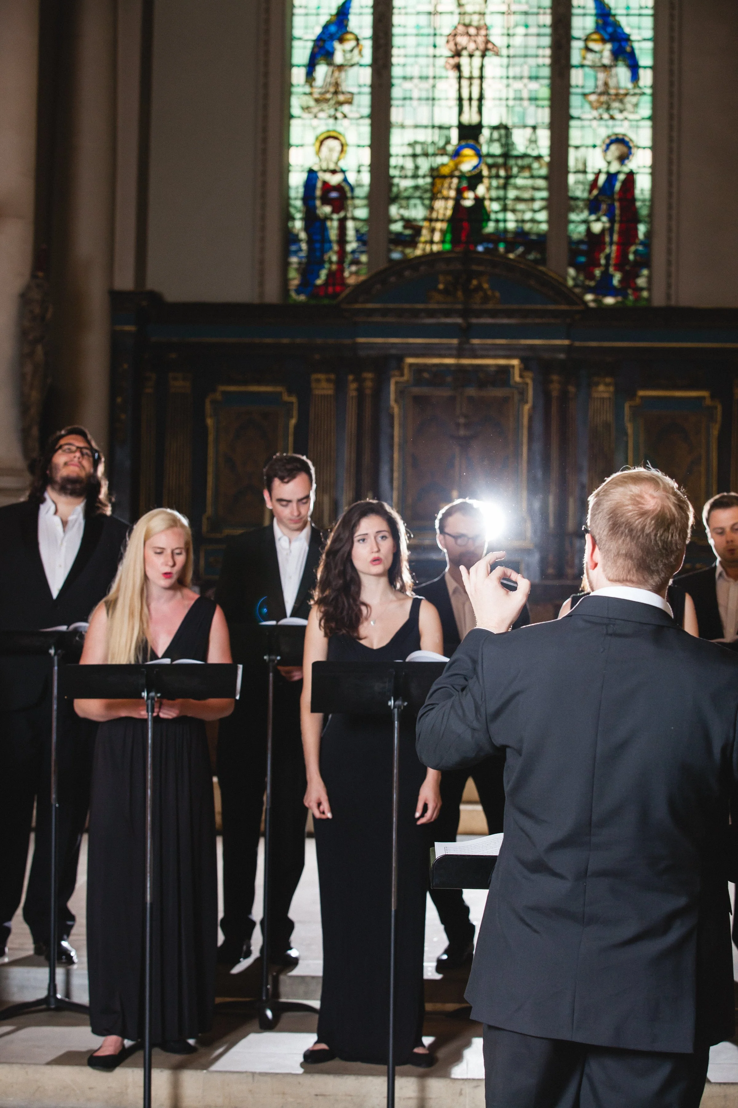 A choir performing inside a church with stained glass windows above, including religious figures and a cross.