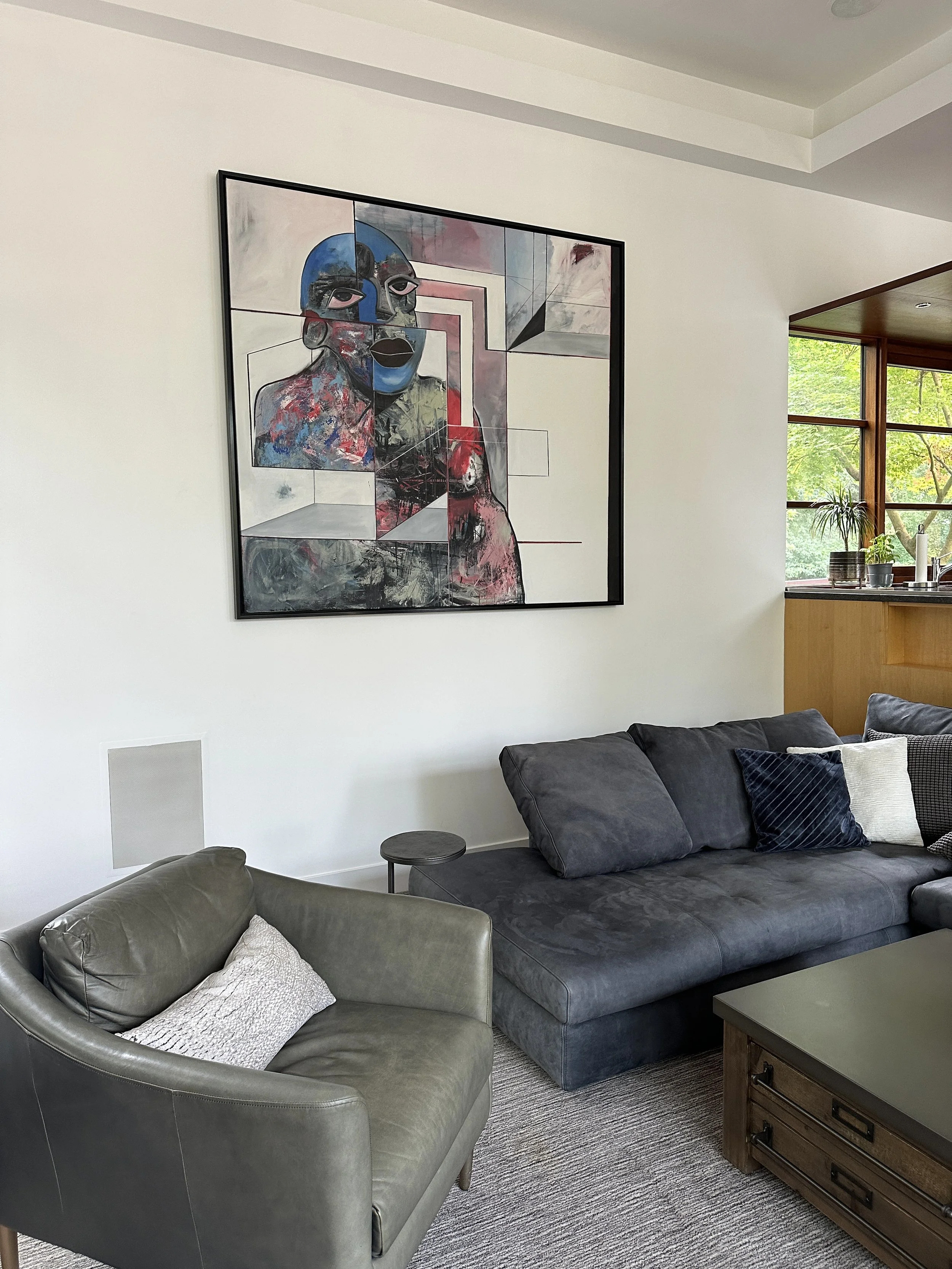 Modern living room with abstract art on wall, gray sectional sofa, leather armchair, coffee table, and large windows.