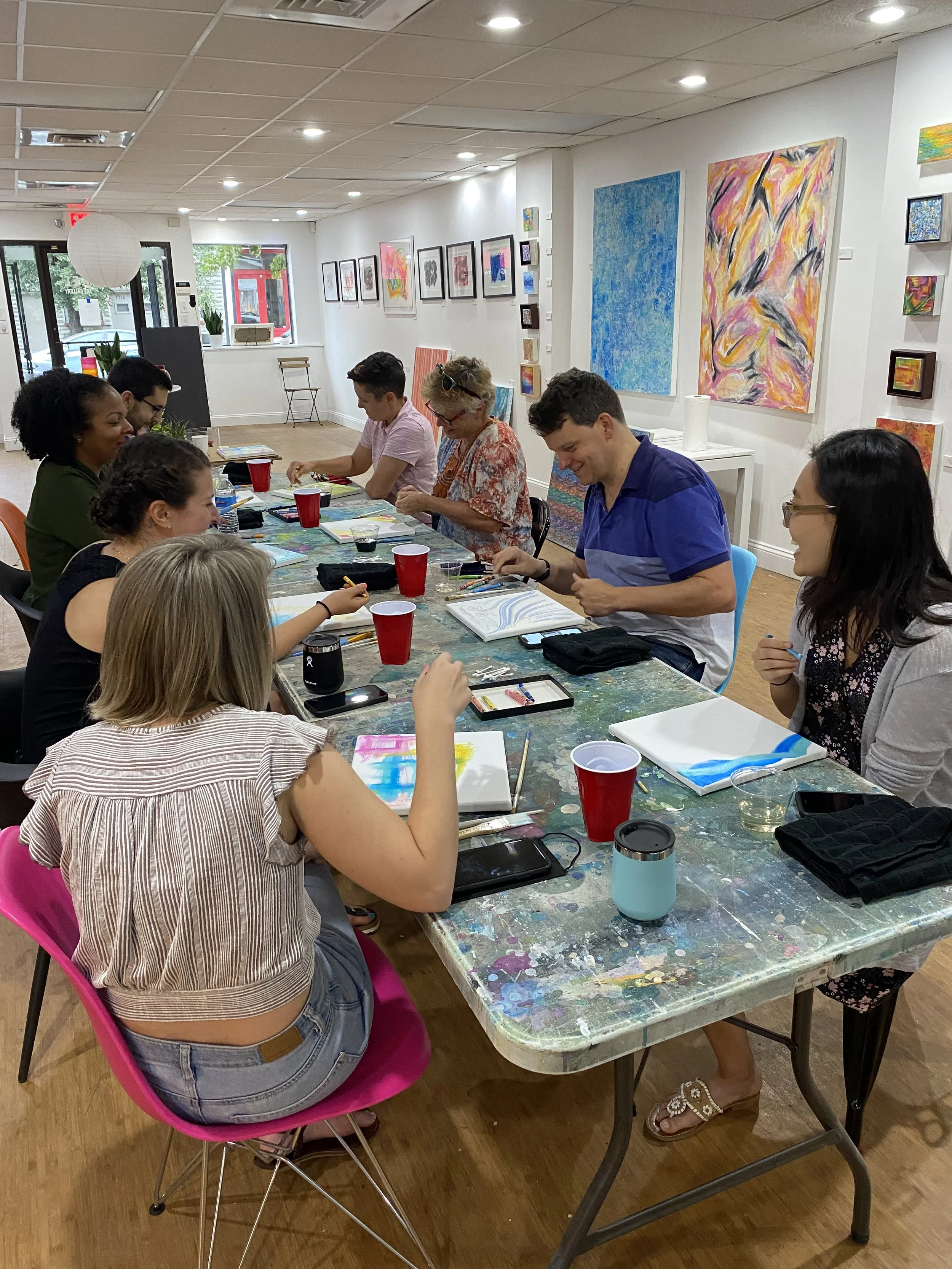 A group of people in an art studio sitting around a table engaged in a painting class. The room has colorful paintings on the walls and art supplies on the table. Some individuals are painting with watercolor sets and brushes. The setting is casual and creative.