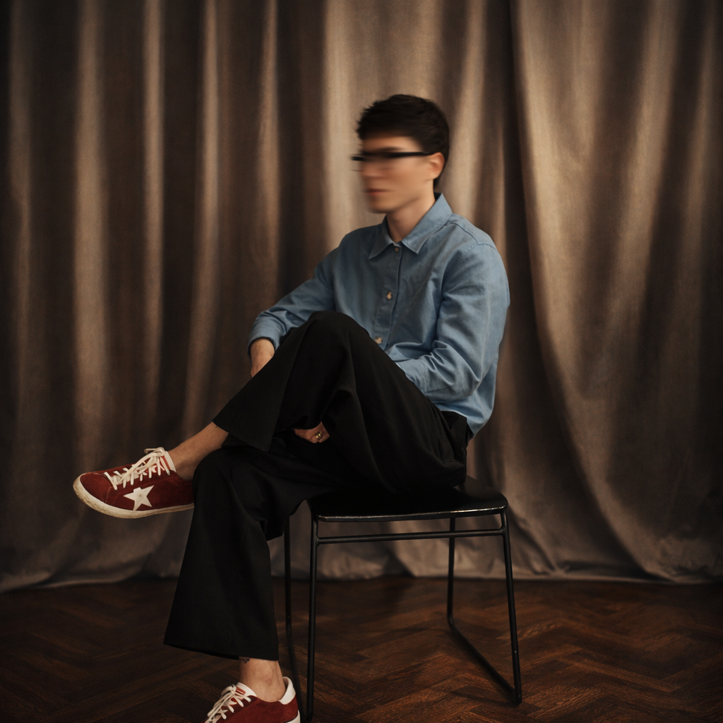 A person with short dark hair, wearing glasses, a blue shirt, black pants, and red sneakers with white star and laces, sitting on a black chair in front of beige curtains.