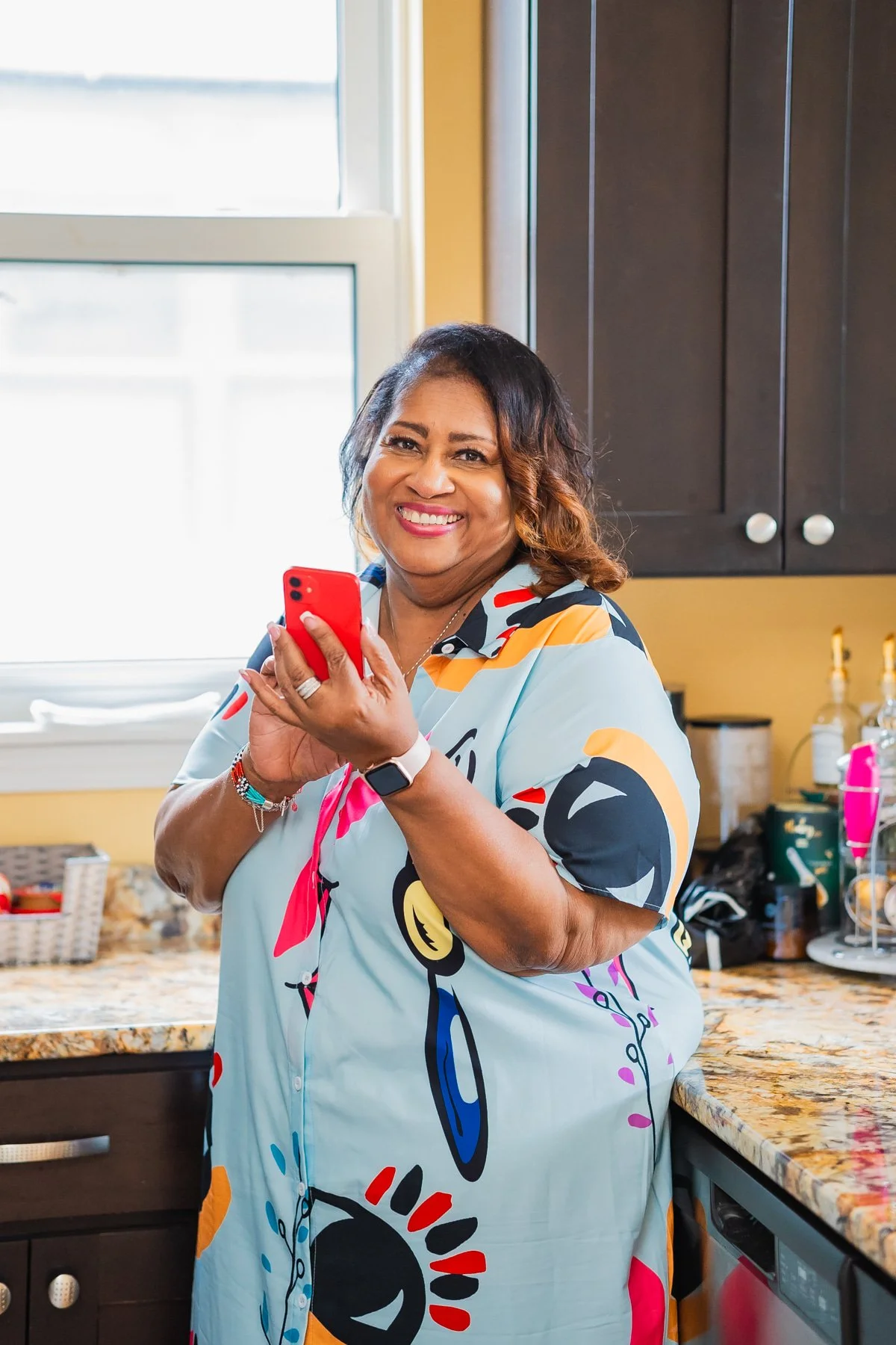 A smiling woman in a colorful dress holding a red phone in a kitchen.