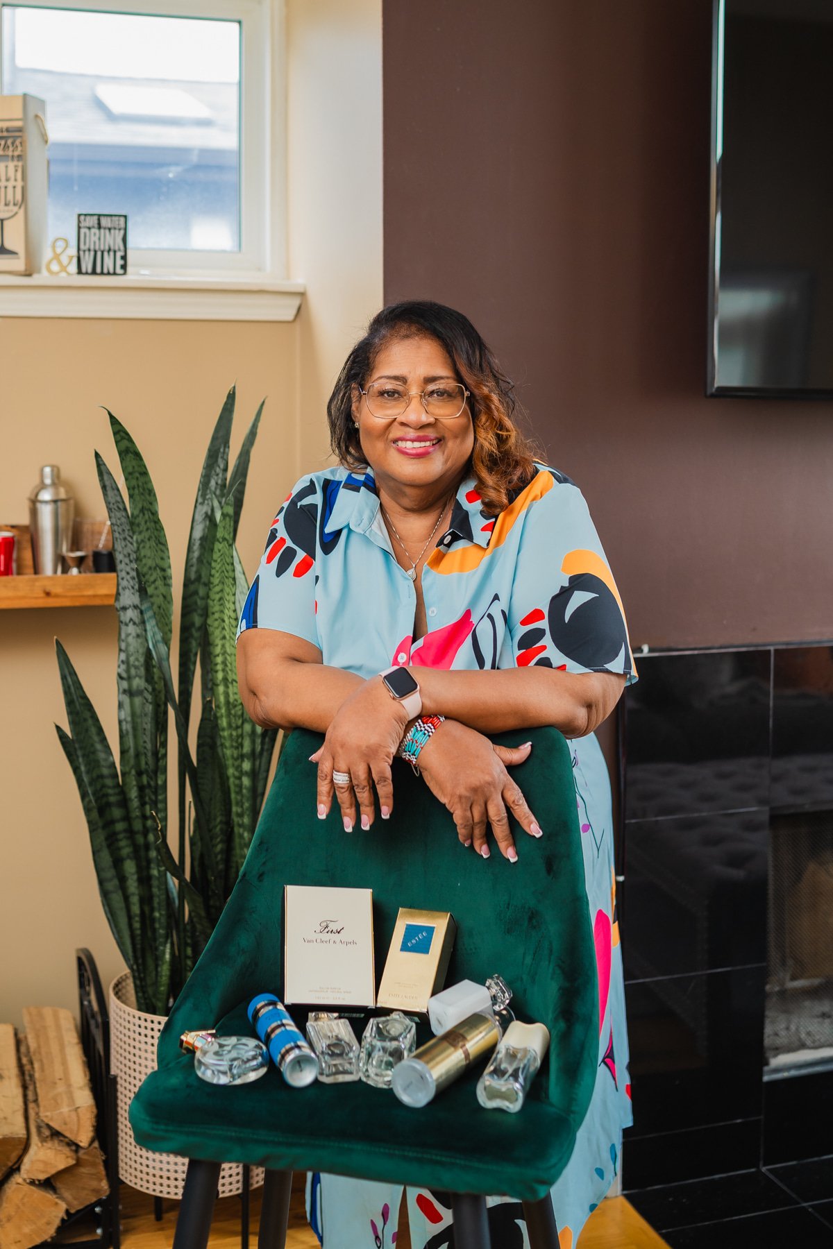 Smiling woman with glasses leaning on a green chair displaying various perfumes and boxes, standing in a room with a fireplace and plants.