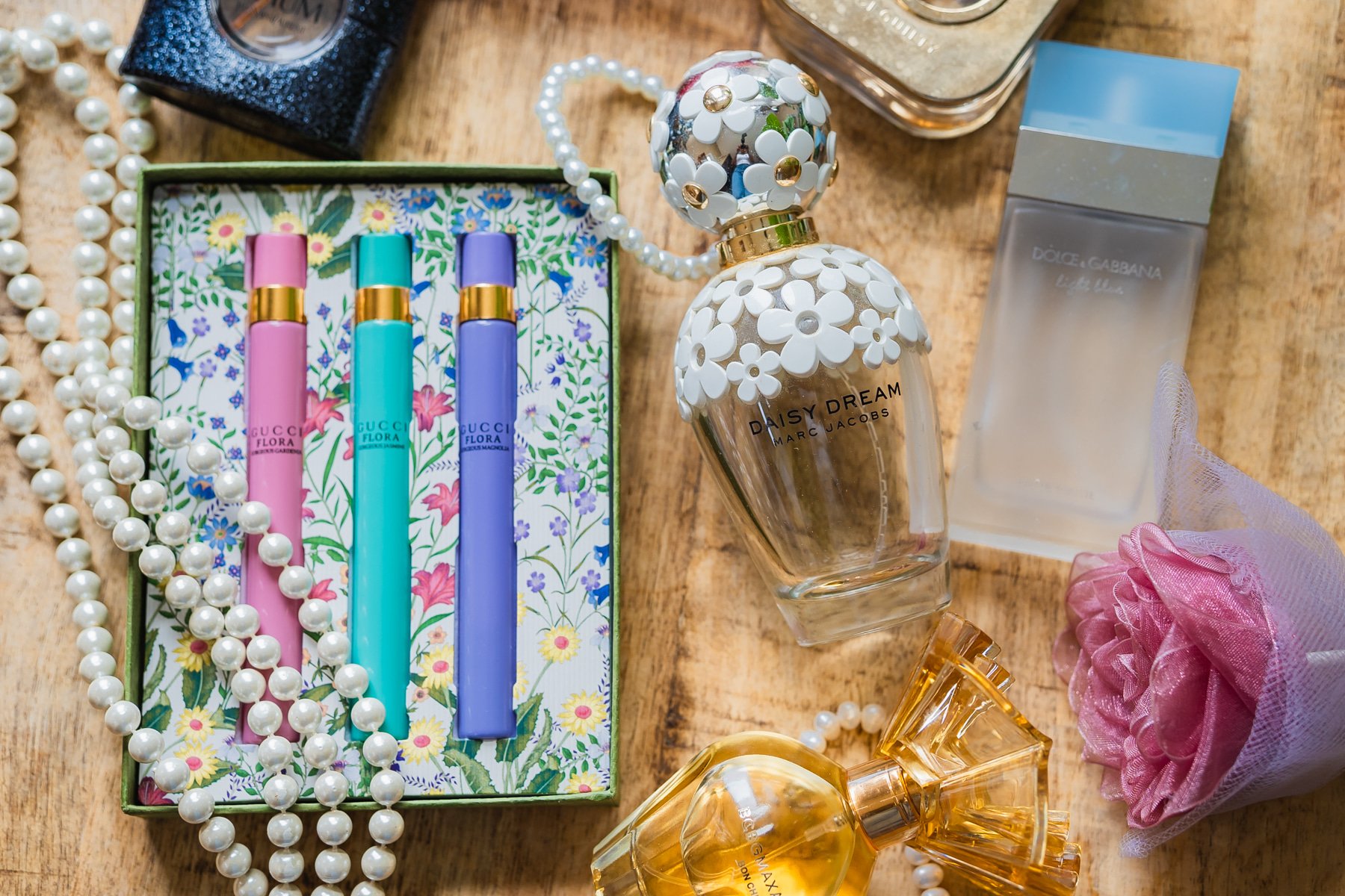 A collection of perfume bottles including Daisy Dream by Marc Jacobs and Gucci Flora, placed on a wooden surface with a pearl necklace and pink decorative flower.